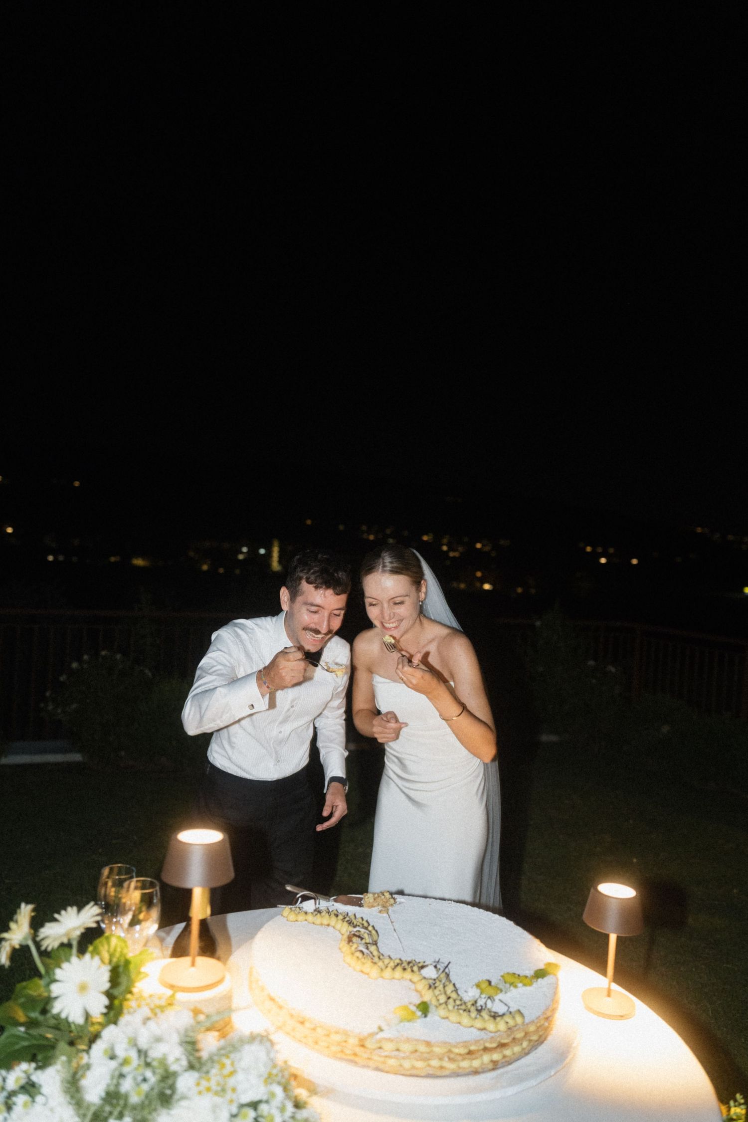 Bride and groom laughing while cutting their decorated wedding cake illuminated by small table lamps at night.
