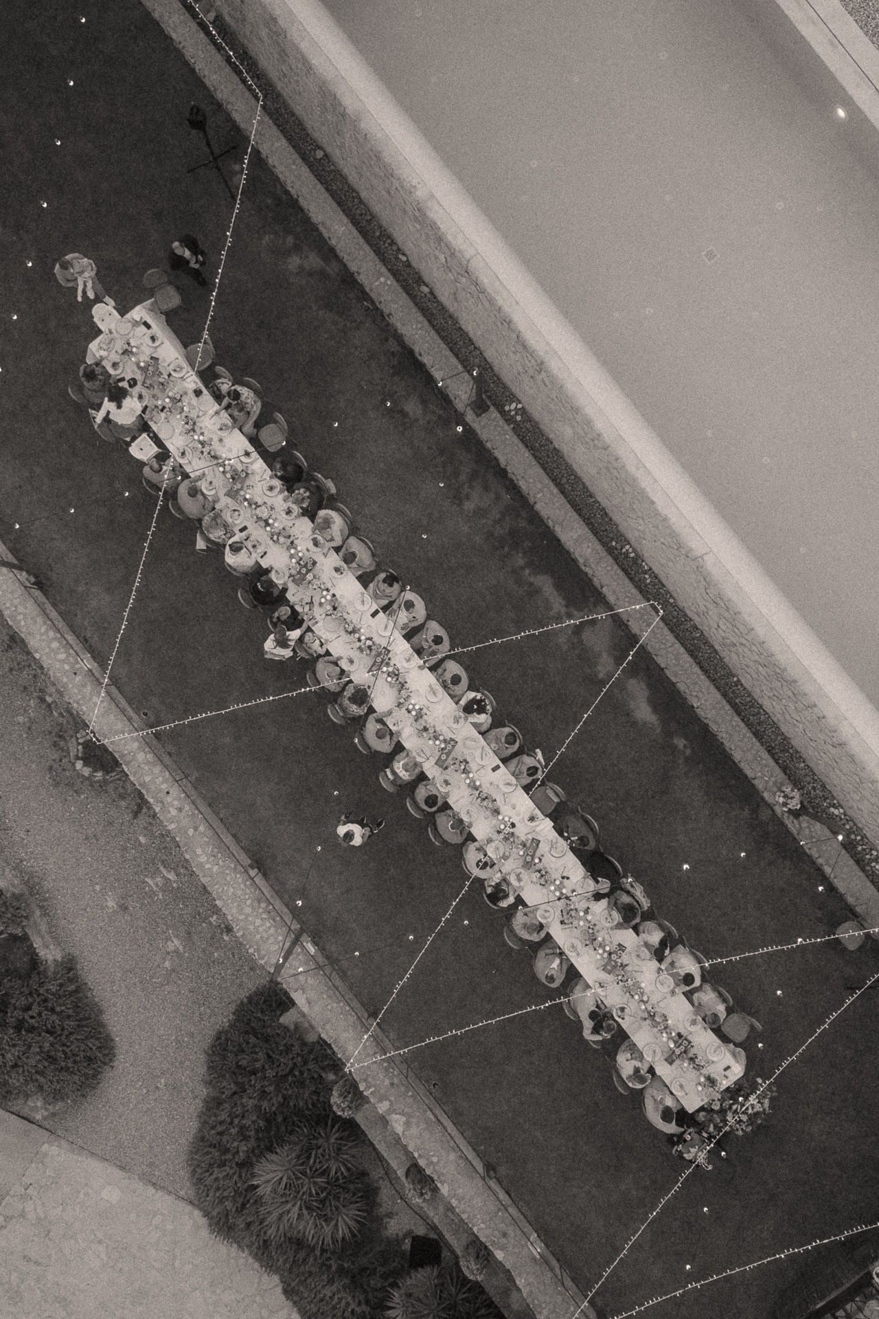 Aerial black and white photo of a long elegant dining table set for an outdoor event with guests seated on both sides.