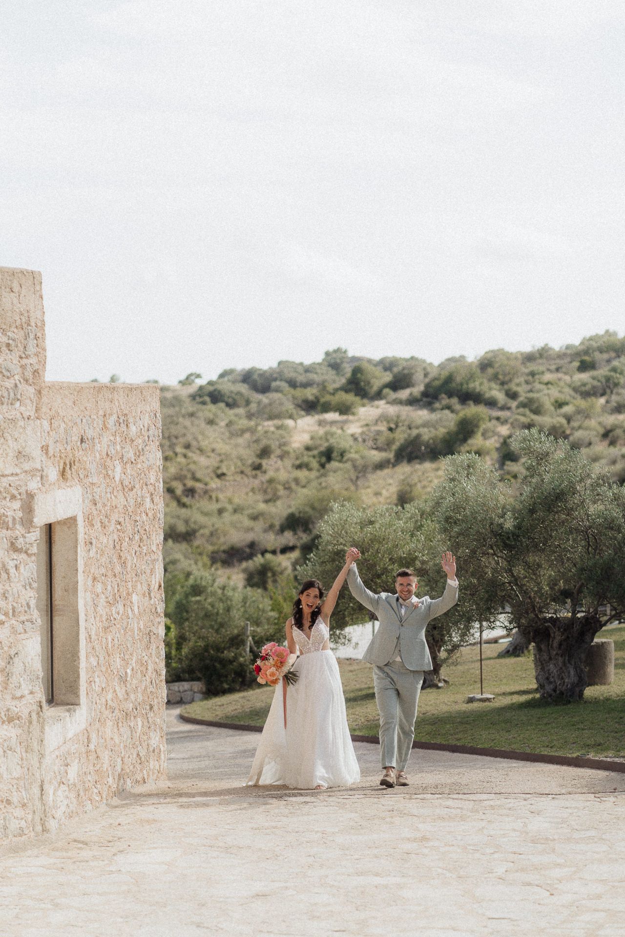 Joyful newlyweds celebrate outdoors at a stone venue with lush green hills, bride holding bouquet in white gown, groom in gray suit.