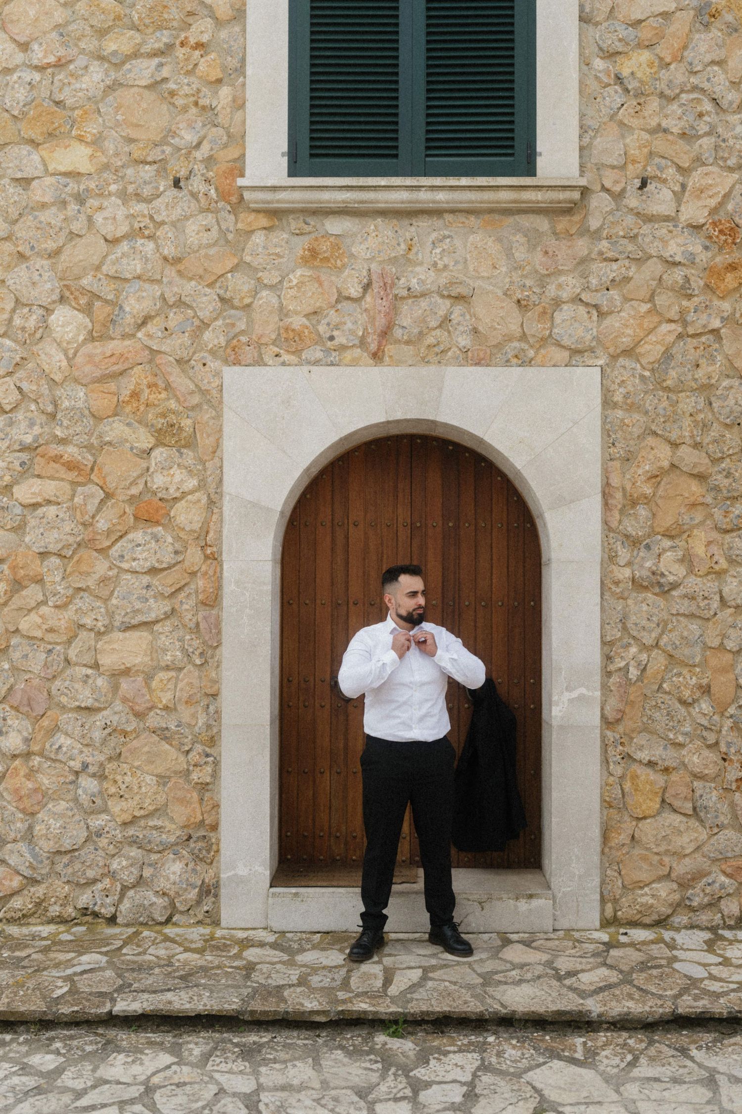 Groom in white shirt adjusting tie standing in front of arched wooden door on stone wall.