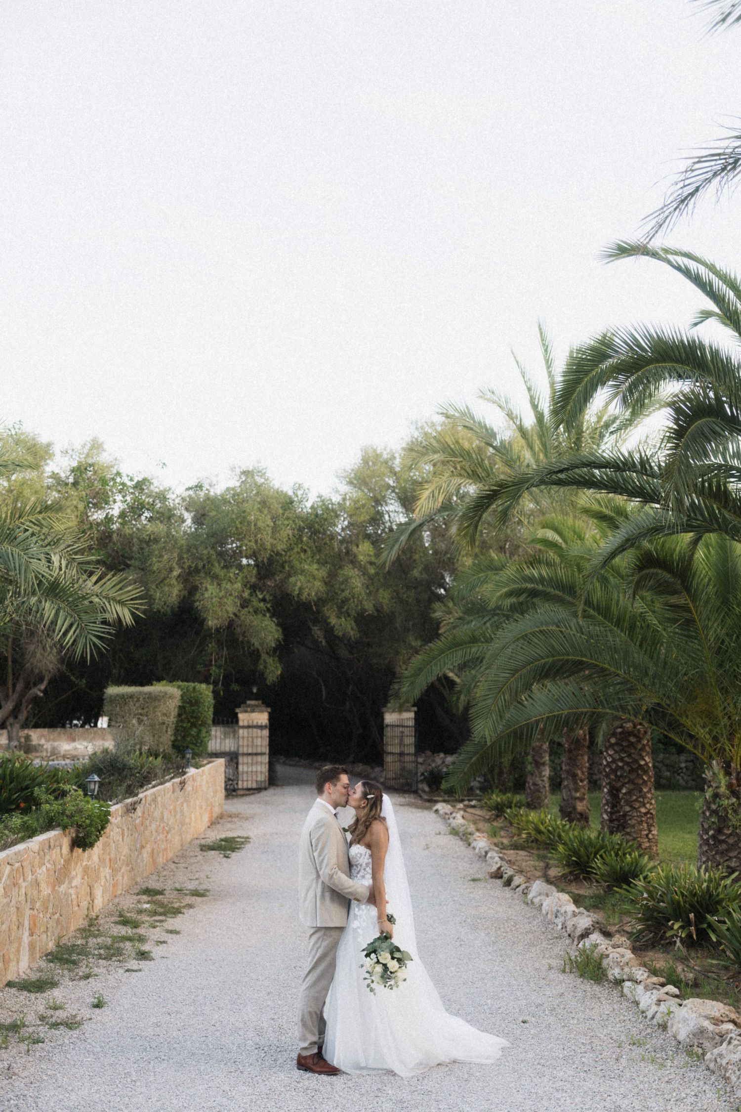 Bride and groom sharing a tender kiss on a gravel path surrounded by tall green palm trees.
