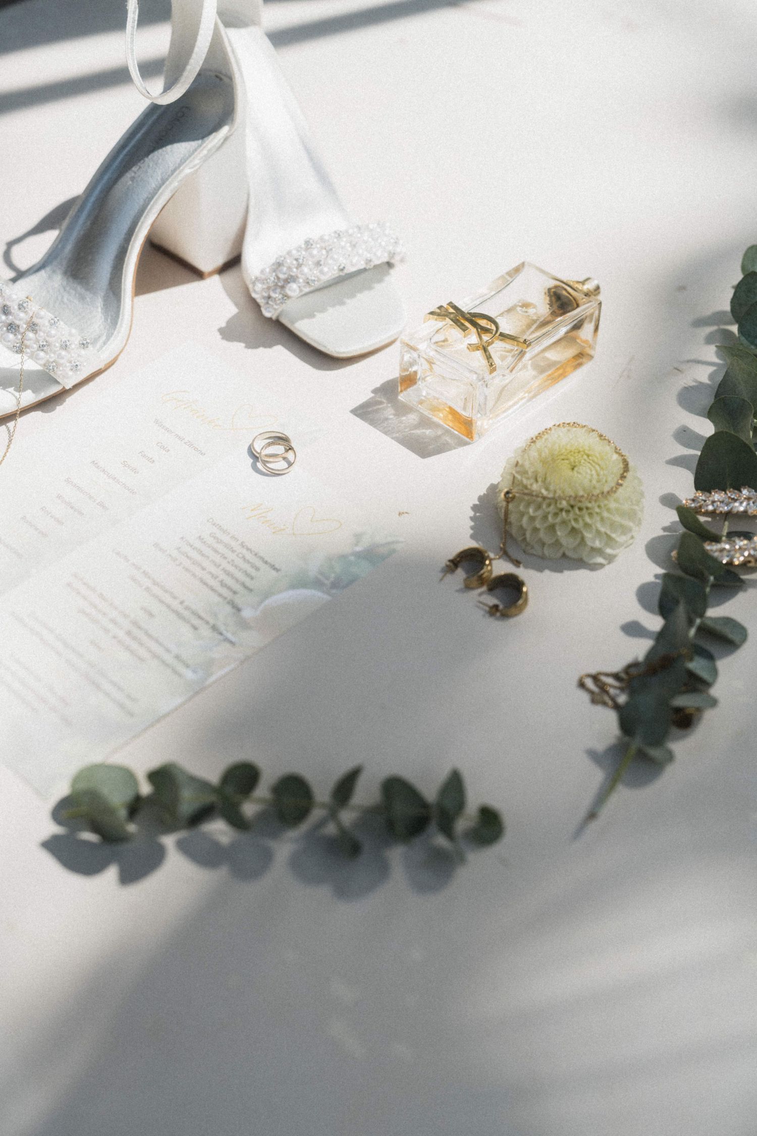 Bridal accessories flat lay with white heels, gold rings, perfume bottle, and green eucalyptus sprigs.