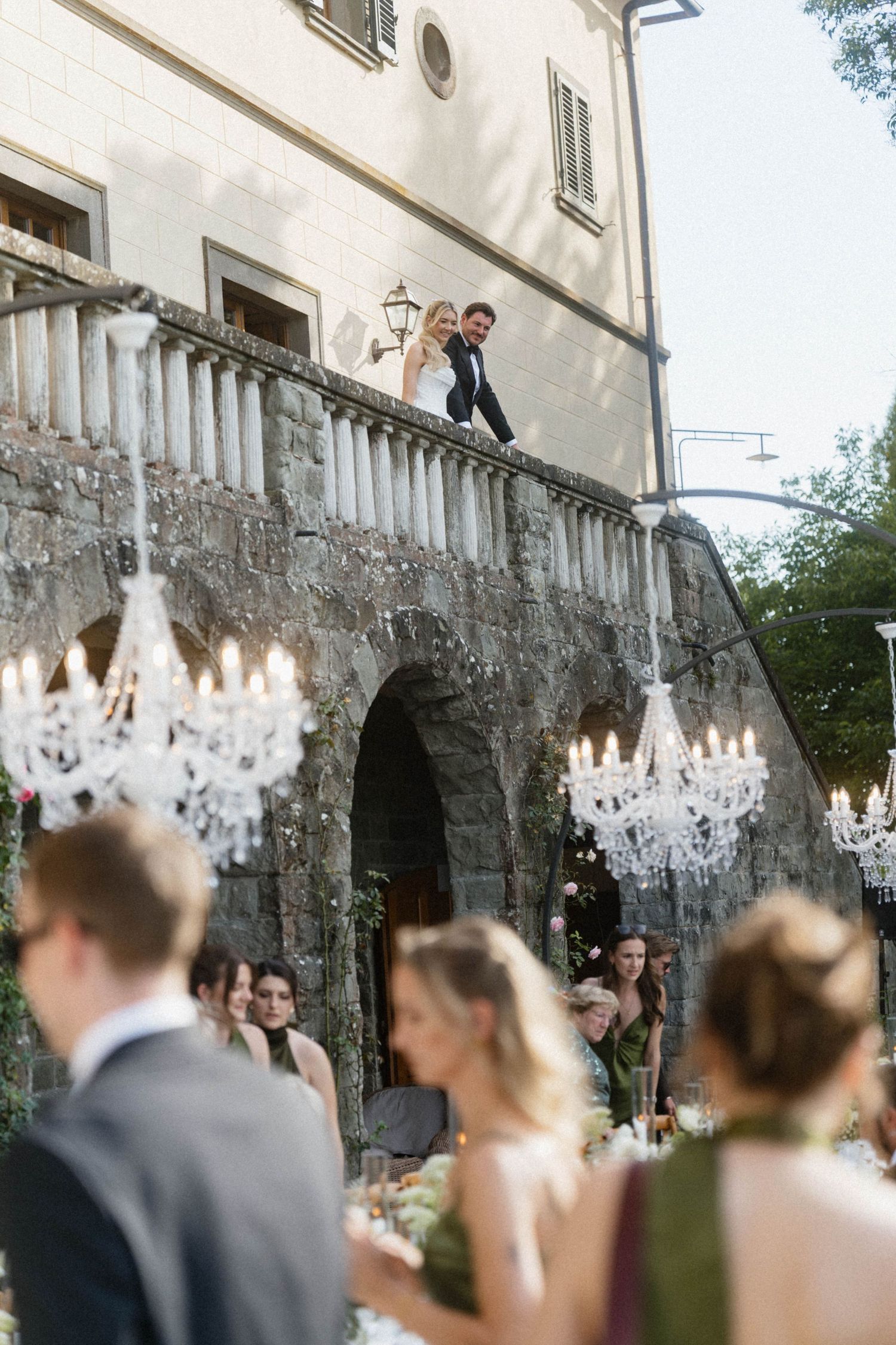 Bride and groom stand on stone balcony overlooking elegant outdoor wedding reception with crystal chandeliers.