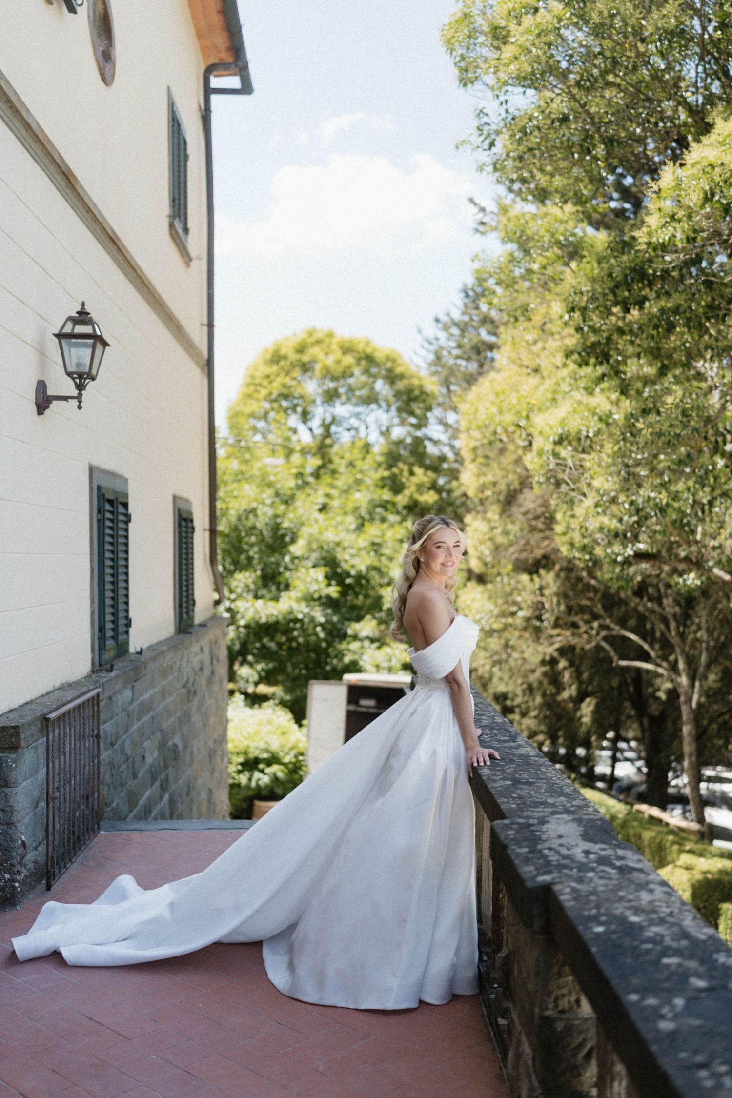 Bride in off-shoulder white wedding gown with train posing on a villa terrace.