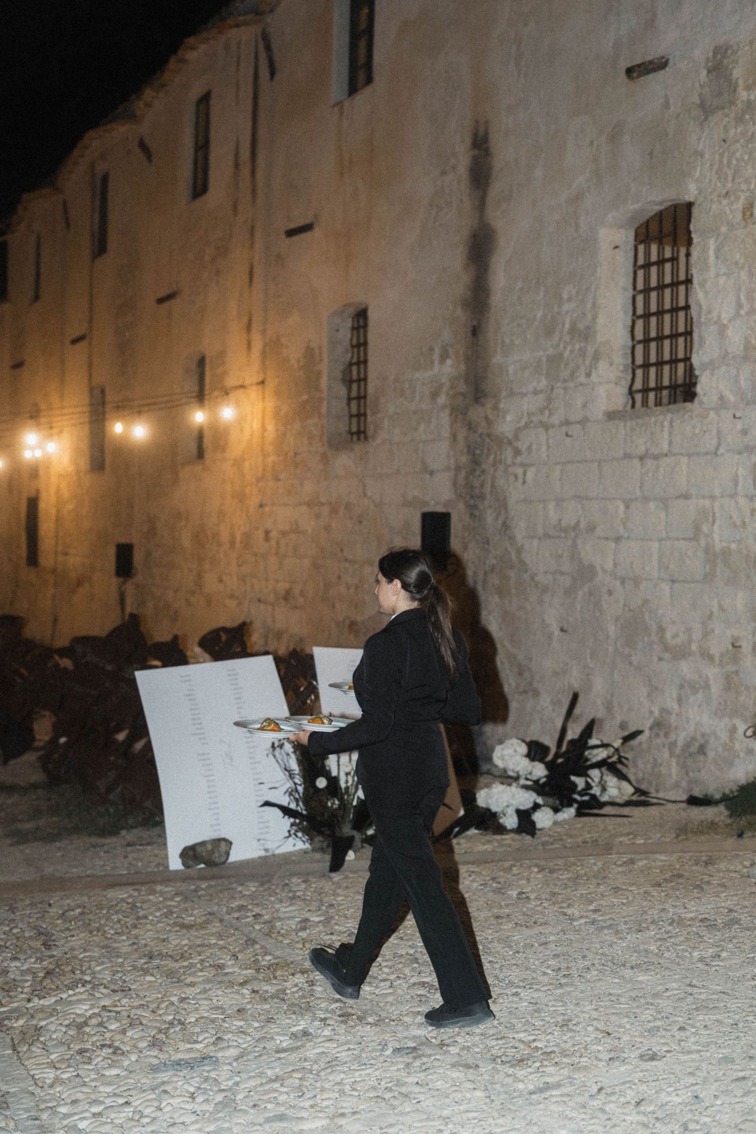 Artist in black carries canvas at night outdoor wedding venue with string lights and stone walls.