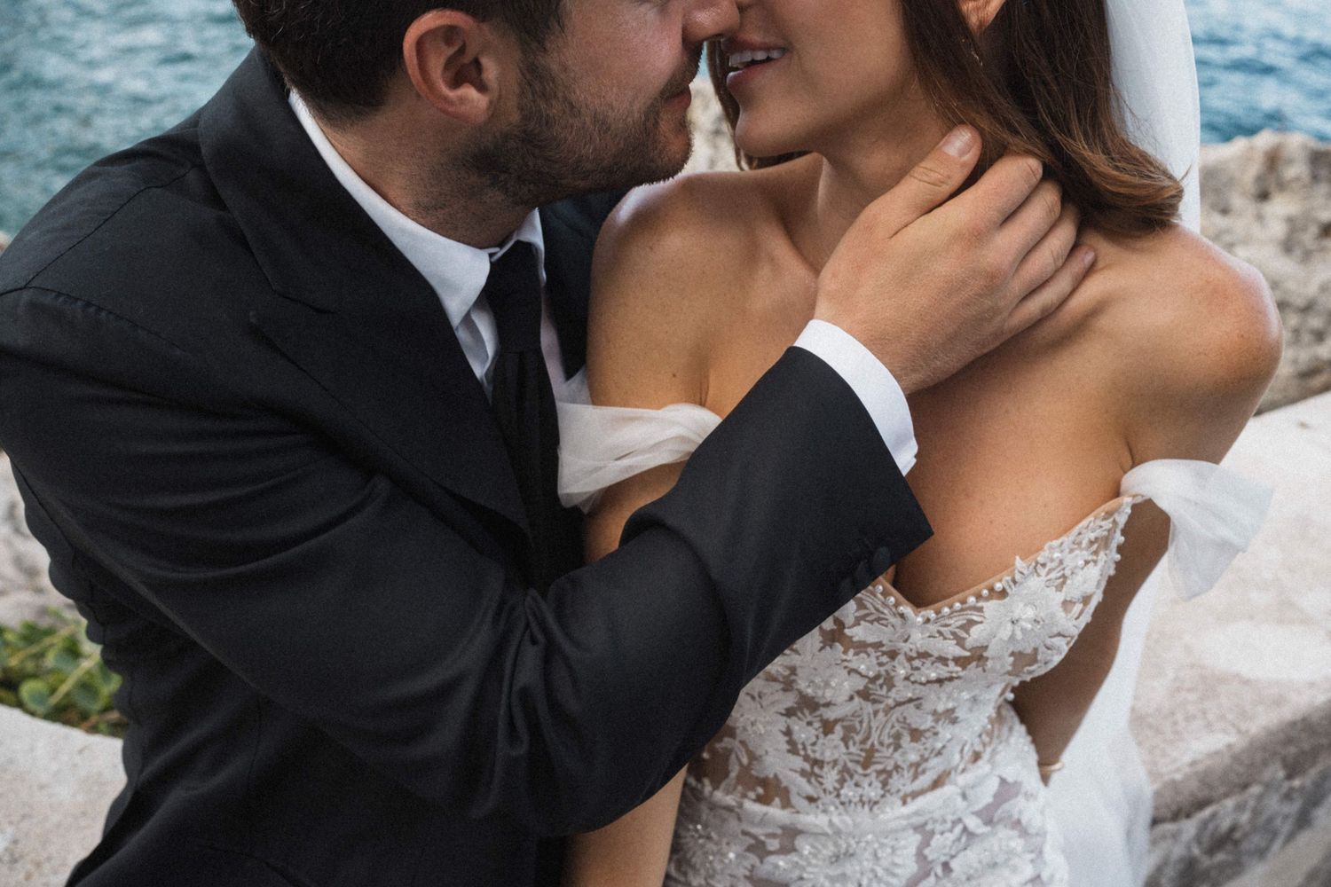Romantic close-up of groom tenderly holding brides neck in lace off-shoulder wedding gown by the sea.