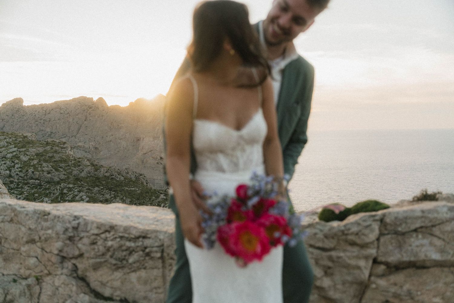 Bride and groom share a romantic sunset moment on a rocky cliff overlooking the sea, bride holding a vibrant pink bouquet.