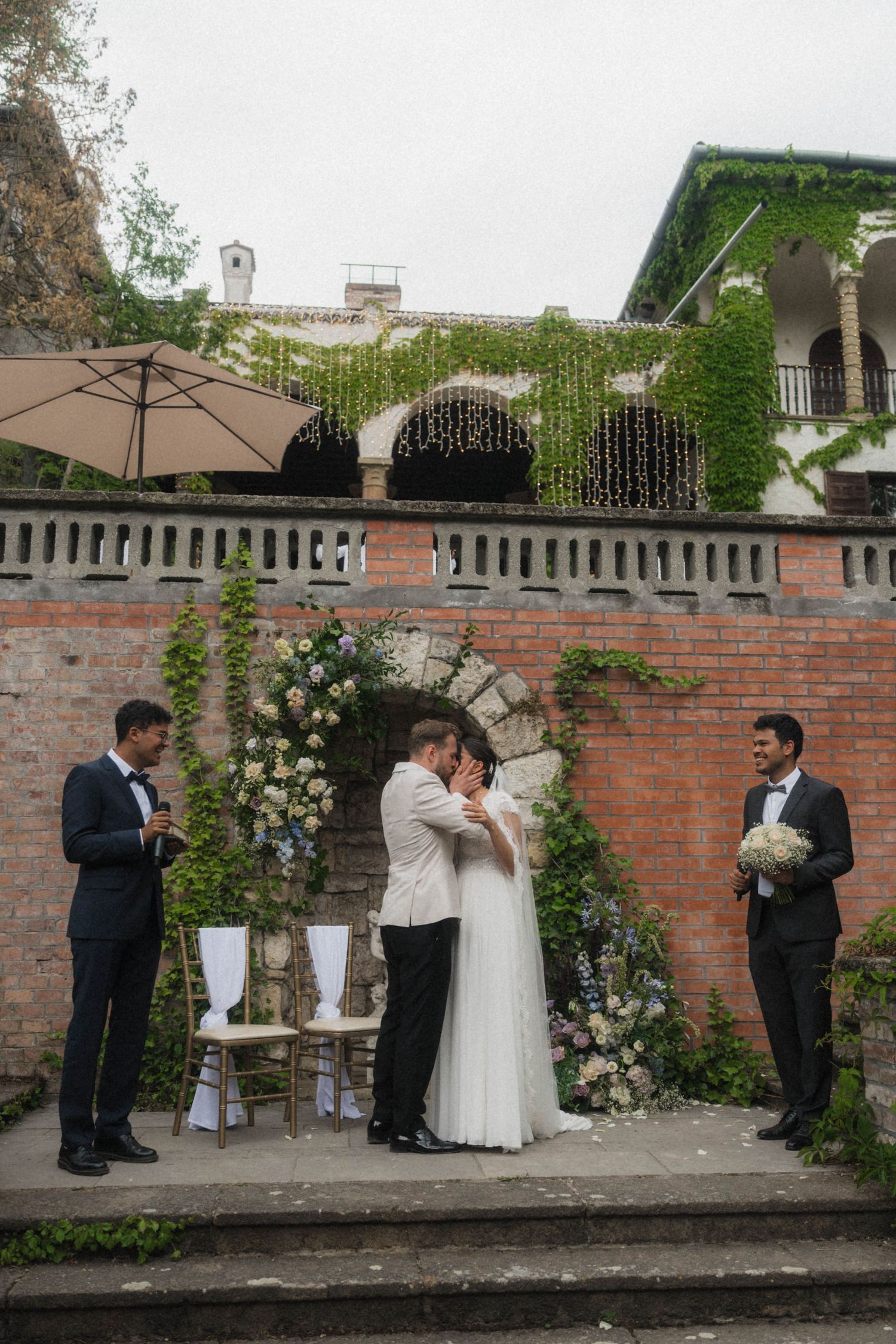 Bride and groom share first kiss at outdoor ceremony with brick arch backdrop.
