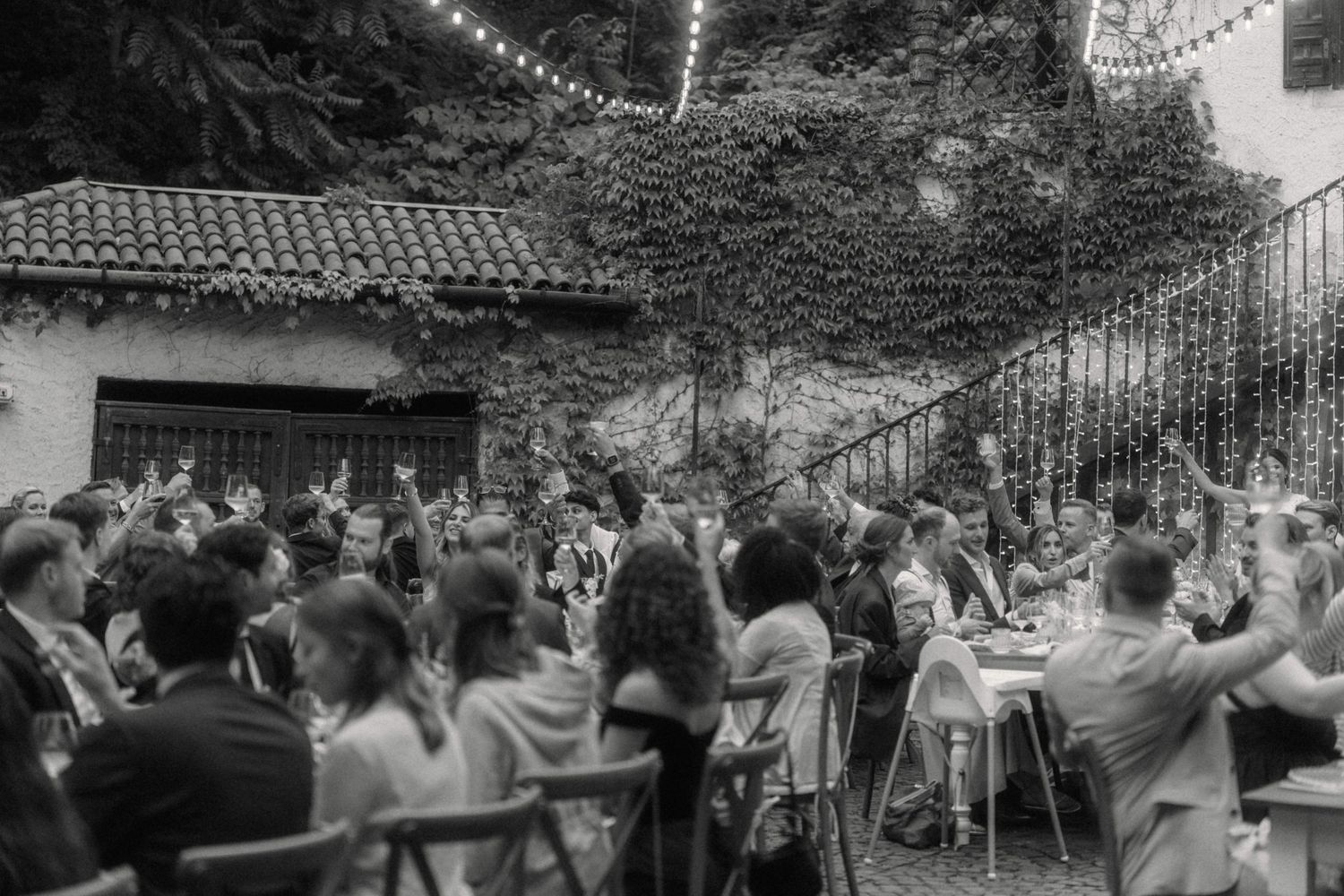 Black and white photo of wedding guests raising glasses in a toast at outdoor garden reception.
