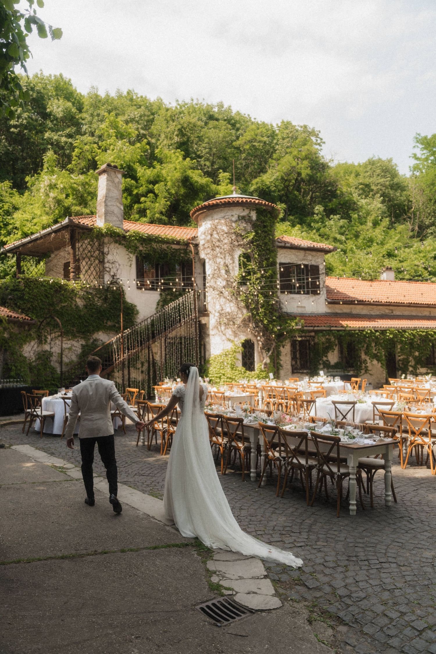 Bride and groom walking toward a rustic stone wedding venue with outdoor reception tables set for dining.