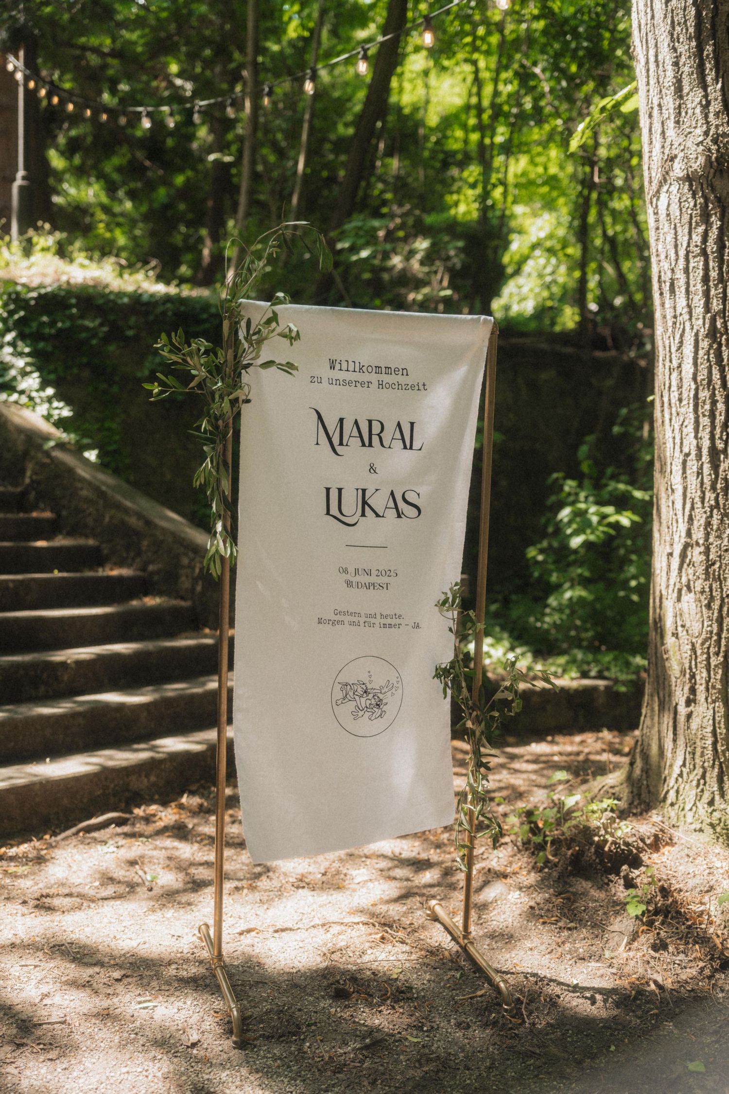 Elegant outdoor wedding welcome sign for Maral and Lukas surrounded by lush greenery and stone steps.