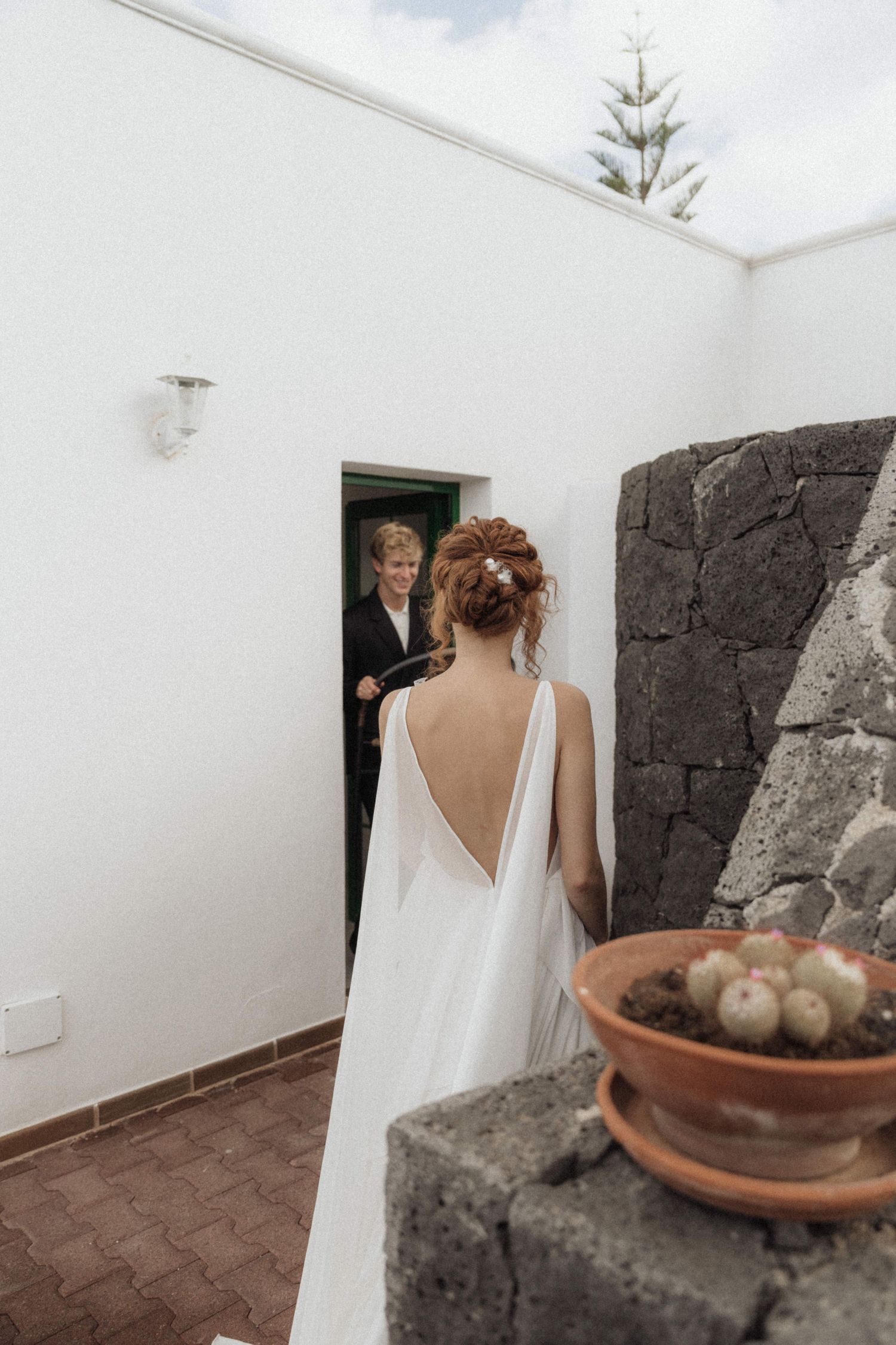Bride in backless white wedding gown faces groom appearing through a doorway in a minimalist white courtyard.
