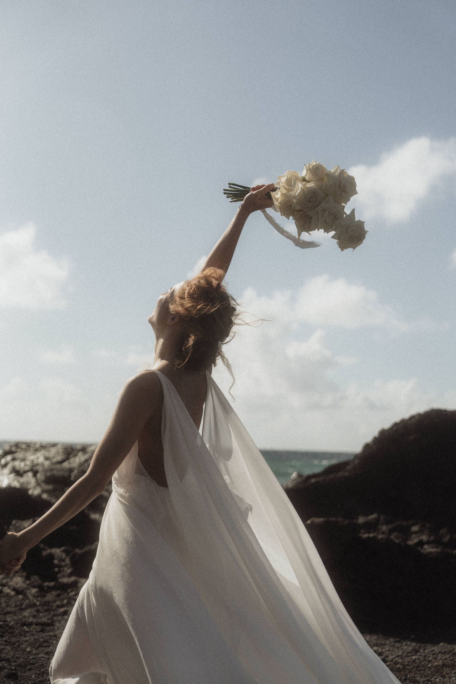 Bride in flowing backless white gown triumphantly raises her bouquet toward the sky at a rocky beach.