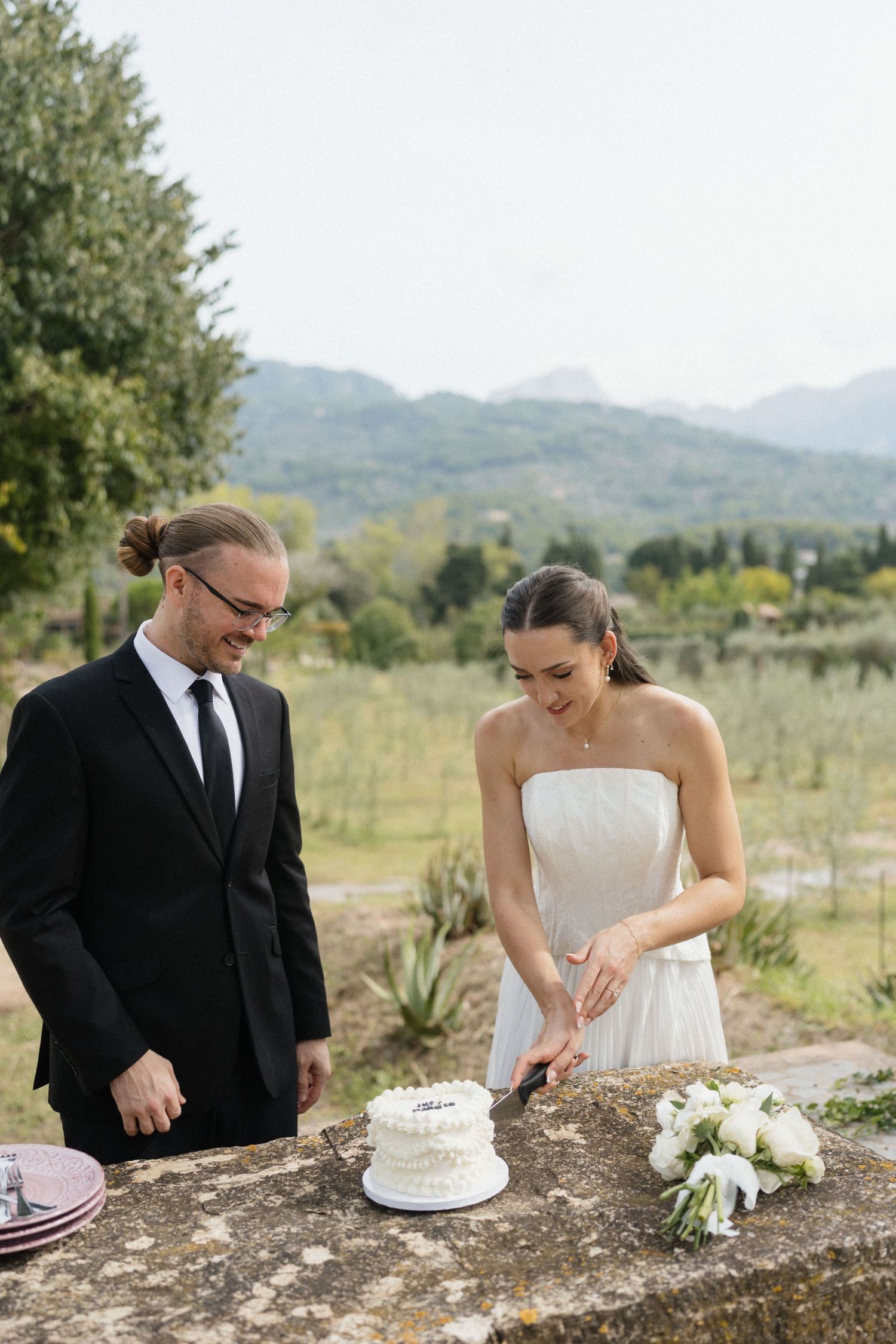 Smiling bride and groom cut a small white wedding cake together on a stone ledge with an olive grove and mountains behind.
