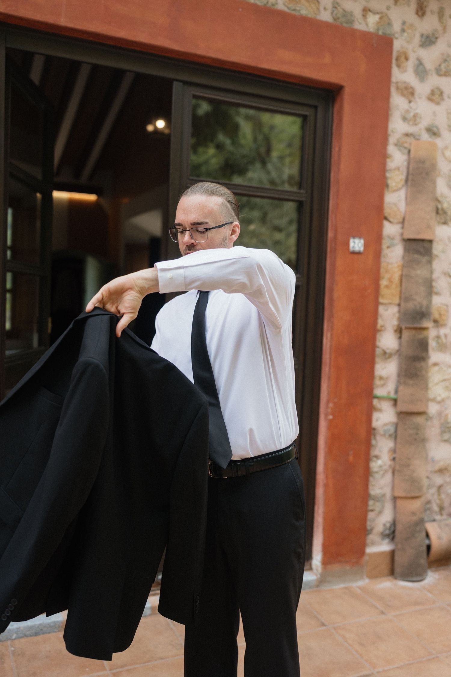 Man in glasses, white dress shirt and black tie puts on a black suit jacket outside a rustic stone building with a wooden door.