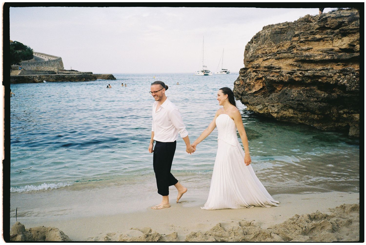 Barefoot couple in white wedding attire holding hands while walking along a sandy beach beside calm turquoise water and rocky cliffs.