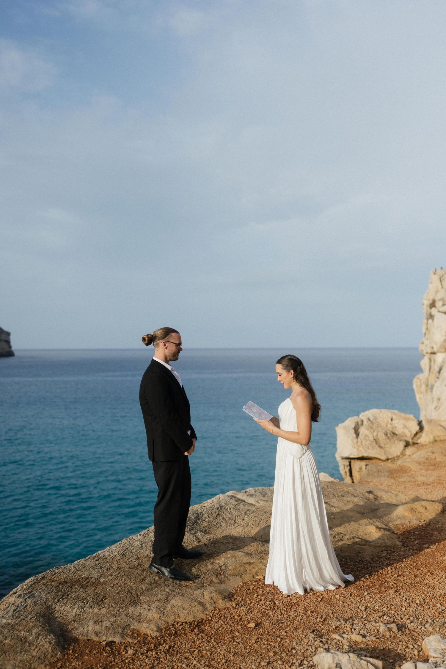 Bride in a white gown and groom in a black suit stand facing each other on a seaside cliff during an elopement ceremony.