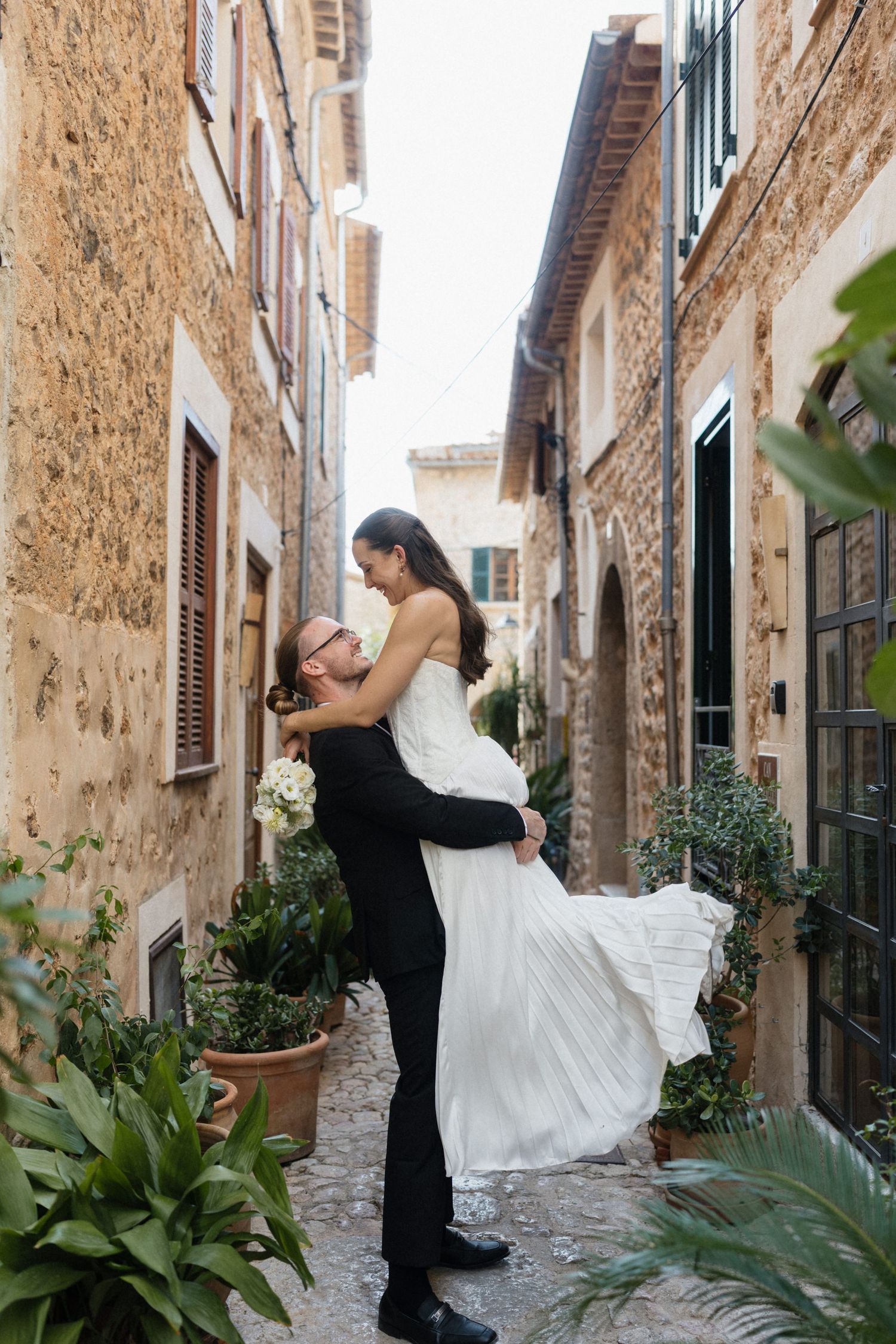 Groom in a black suit lifts a bride in a flowing white gown while she holds a bouquet in a charming stone alley with potted plants.