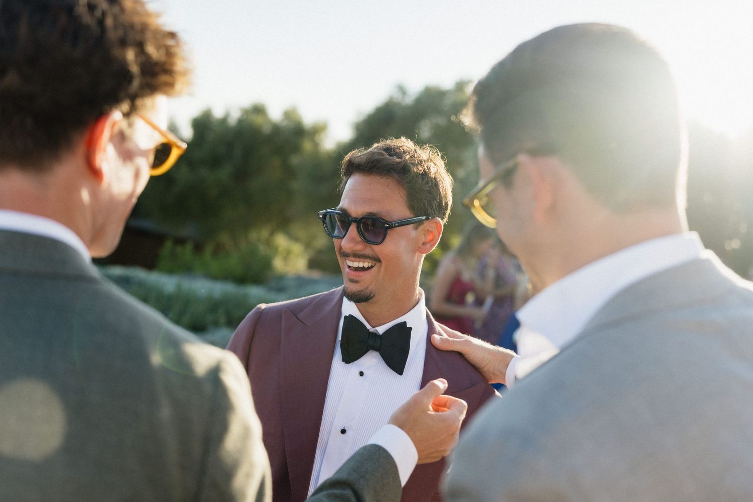 Groom in a burgundy blazer, bow tie and sunglasses laughs with two male guests during a sunny wedding celebration.