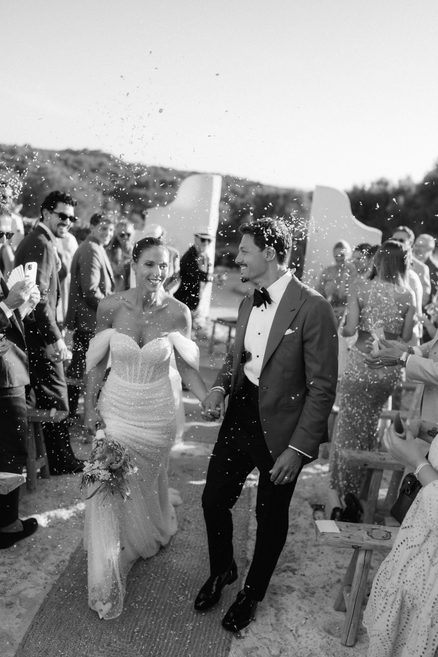 Bride and groom walk hand in hand down the aisle as wedding guests toss confetti at an outdoor ceremony.