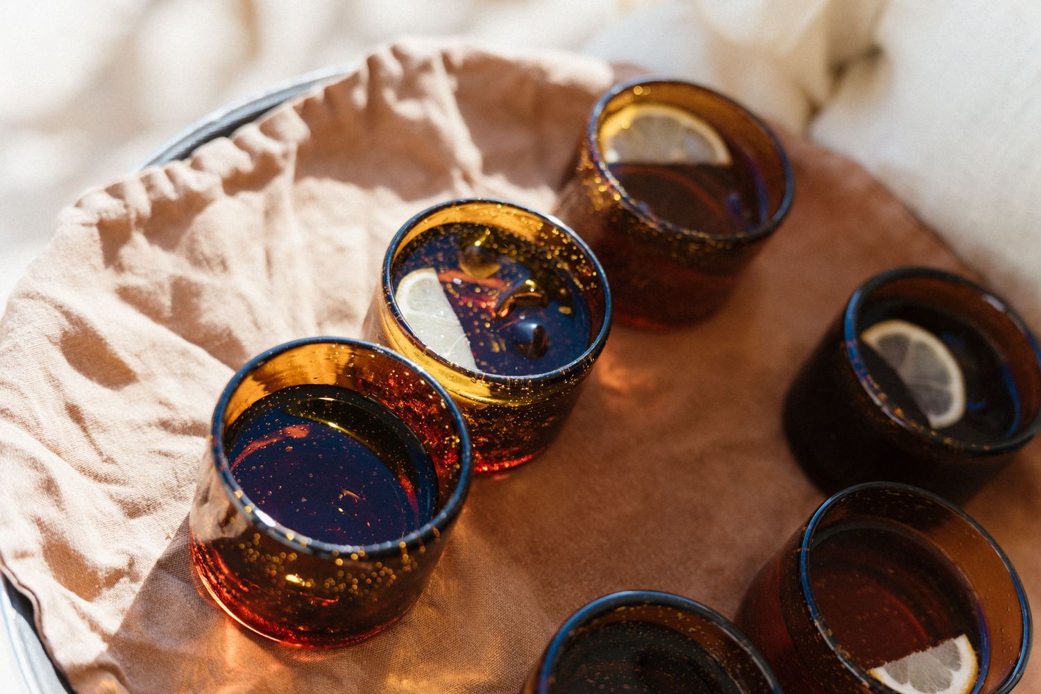 Amber glass tumblers filled with dark iced drinks and lemon slices on a round tray lined with soft linen in warm sunlight.