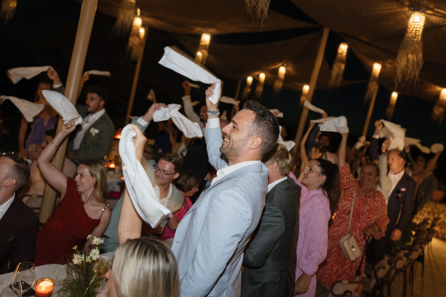 Wedding guests wave white napkins in the air and cheer during a lively evening reception under string lights.