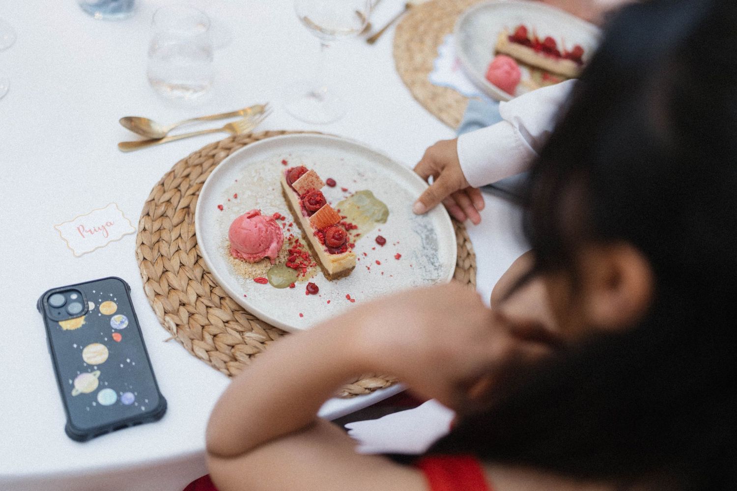 Child reaching for a colorful dessert tart with pink ice cream and fresh berries on a woven placemat at a white table.