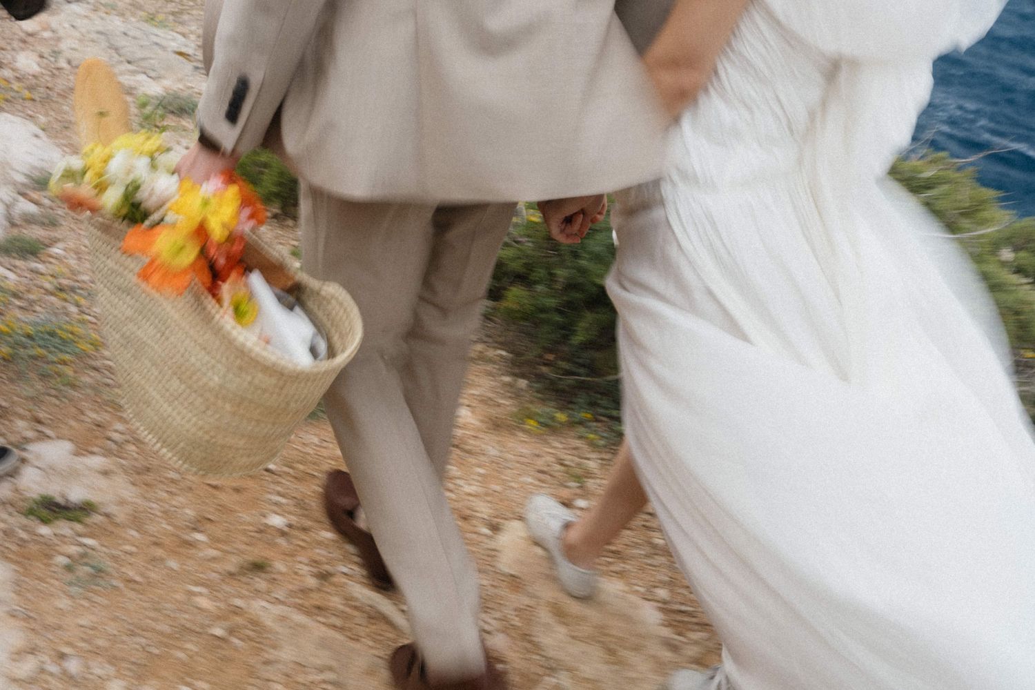 Close-up of bride and groom walking on rocky terrain, groom carrying a wicker picnic basket.