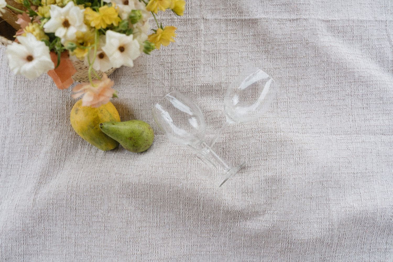 Two crystal wine glasses lying on white linen tablecloth with yellow and white floral arrangement.
