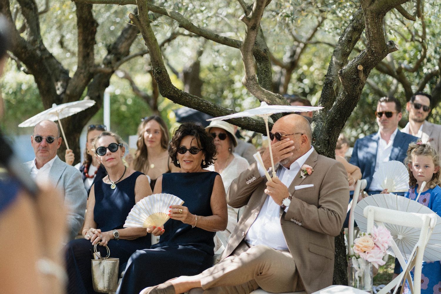 Wedding guests seated outdoors under olive trees, some holding parasols and fans during the ceremony.