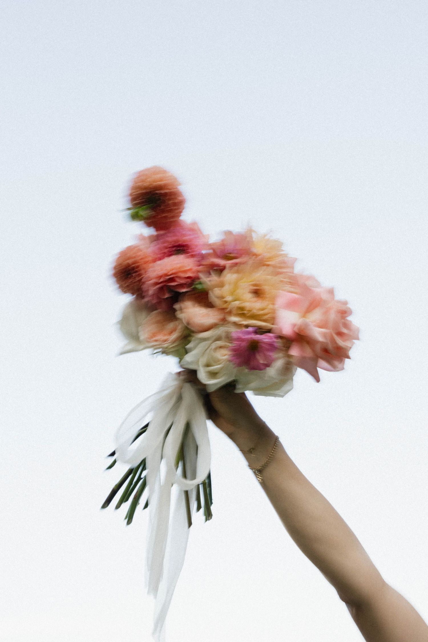 A hand holds a vibrant bouquet of pink, peach, and white flowers with white ribbons against a pale sky.