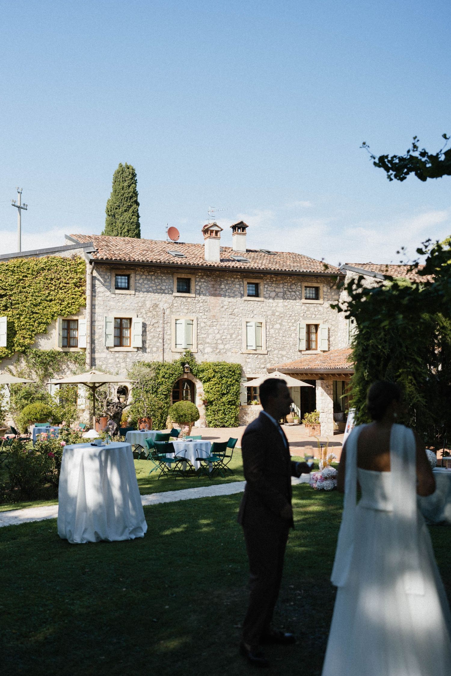 Bride and groom stand in lush garden of an ivy-covered Italian stone villa with elegant wedding reception tables.