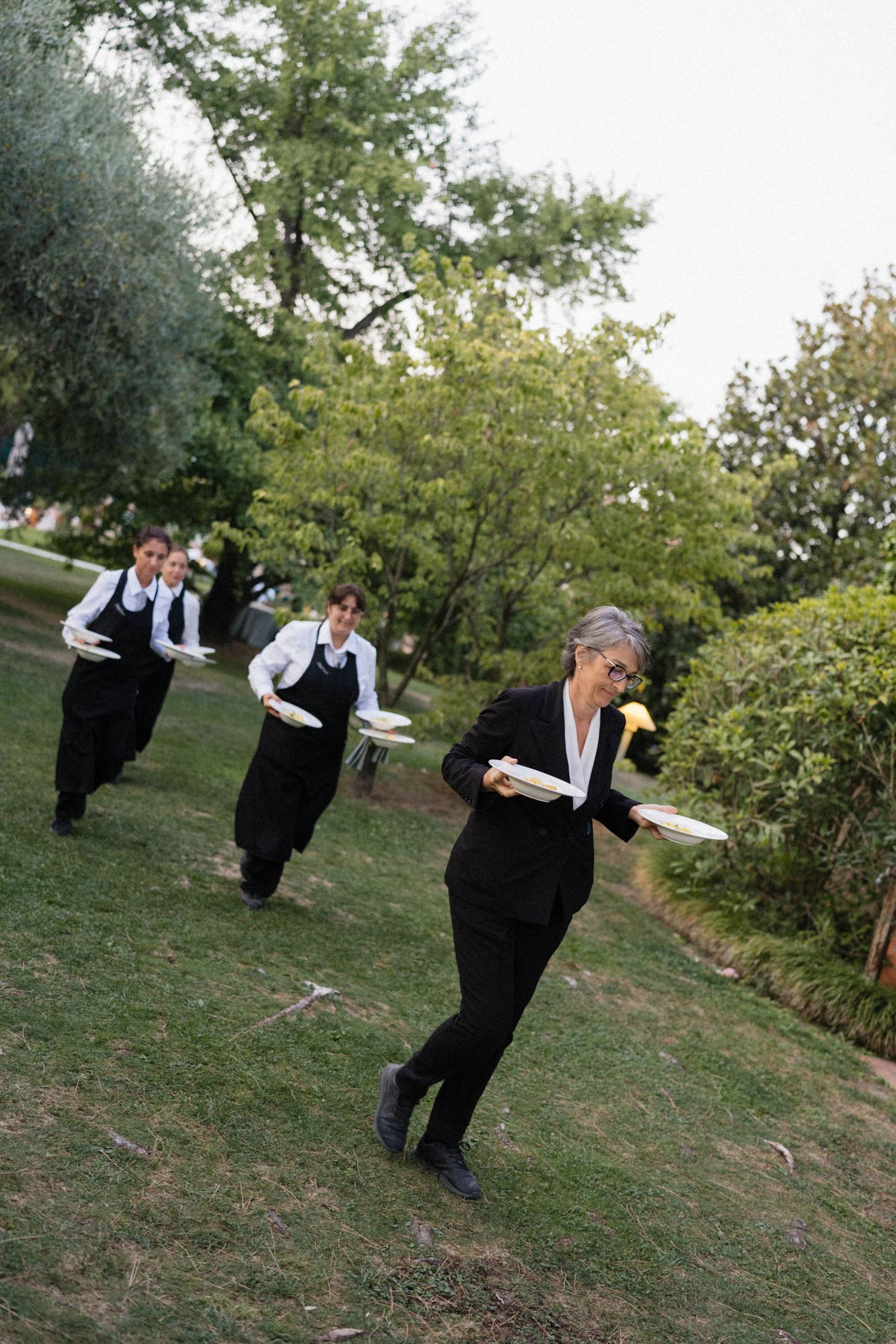 Catering staff in black uniforms carrying white plates across a garden lawn at an outdoor wedding event.