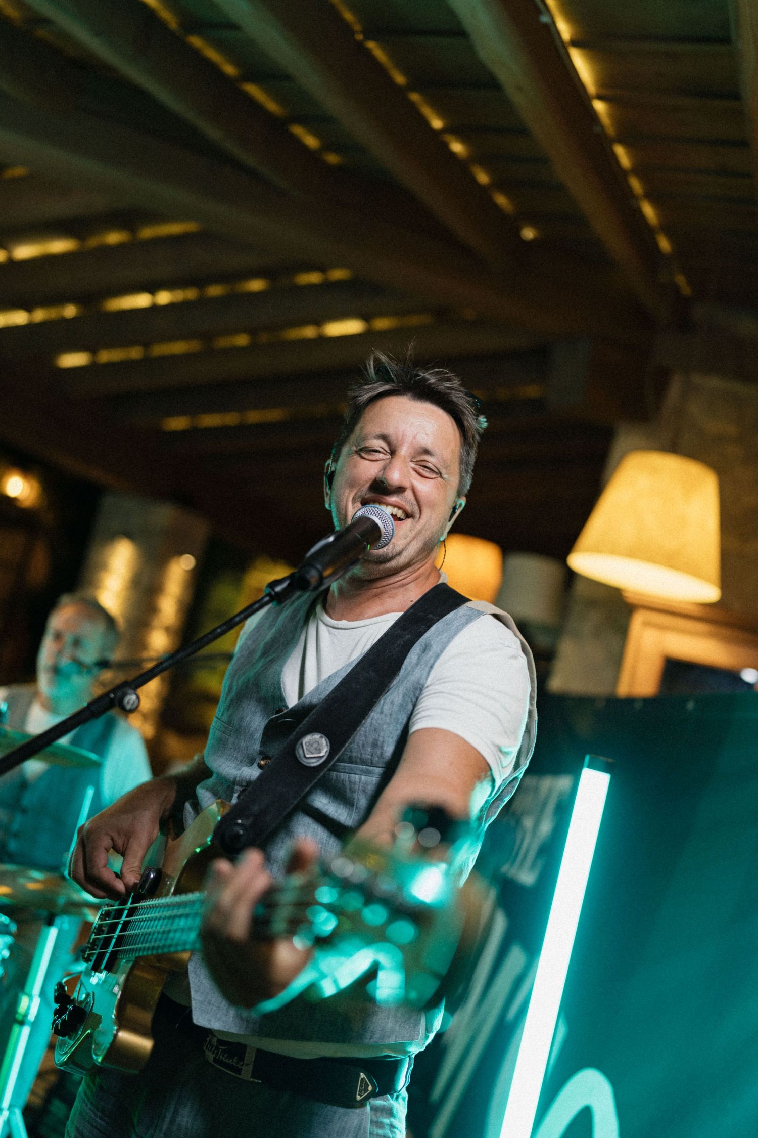 A smiling male musician plays bass guitar and sings into a microphone on stage under warm lights.