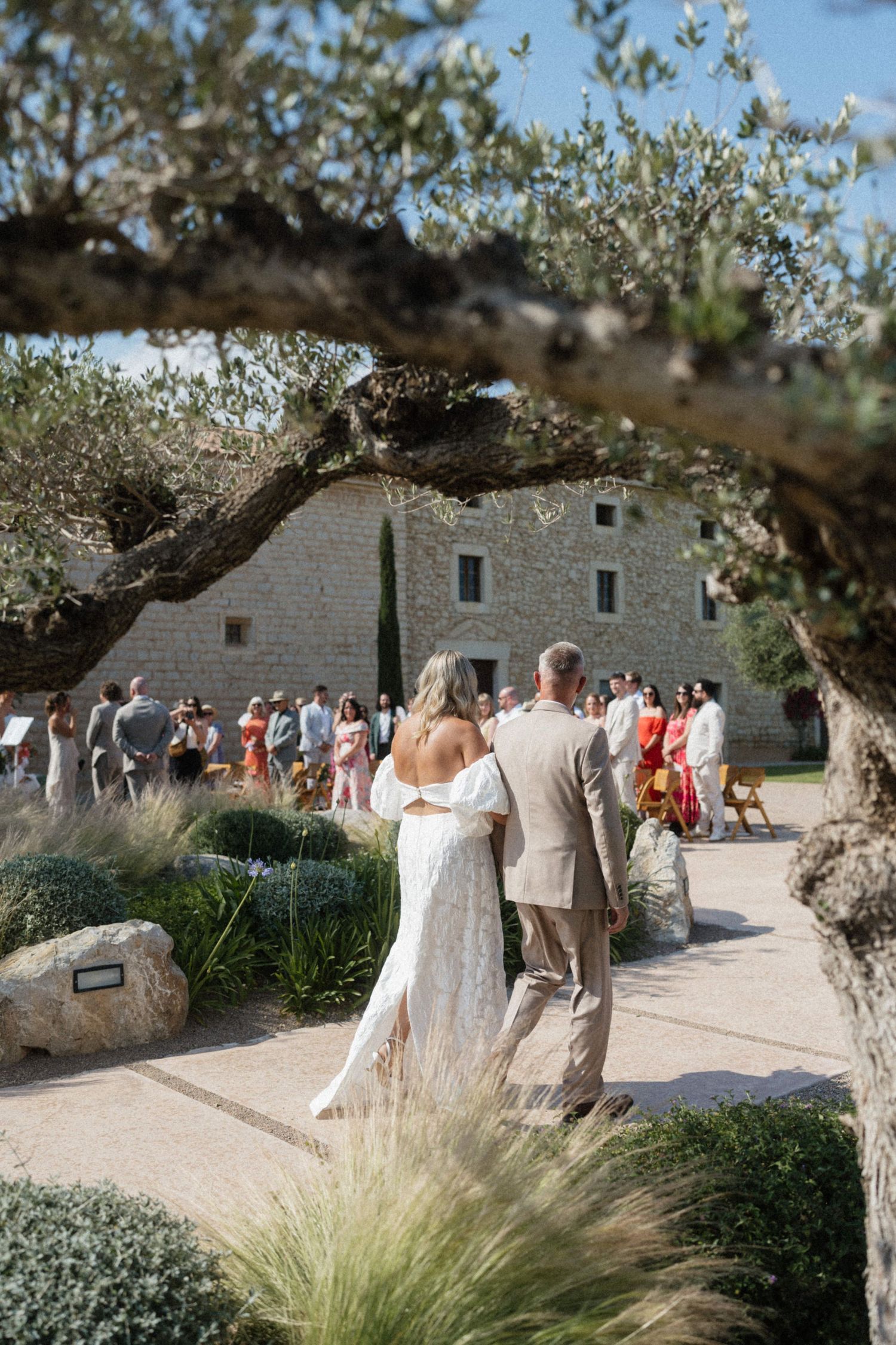 Bride and groom walk toward outdoor wedding guests amid olive trees at a sunlit stone villa venue.