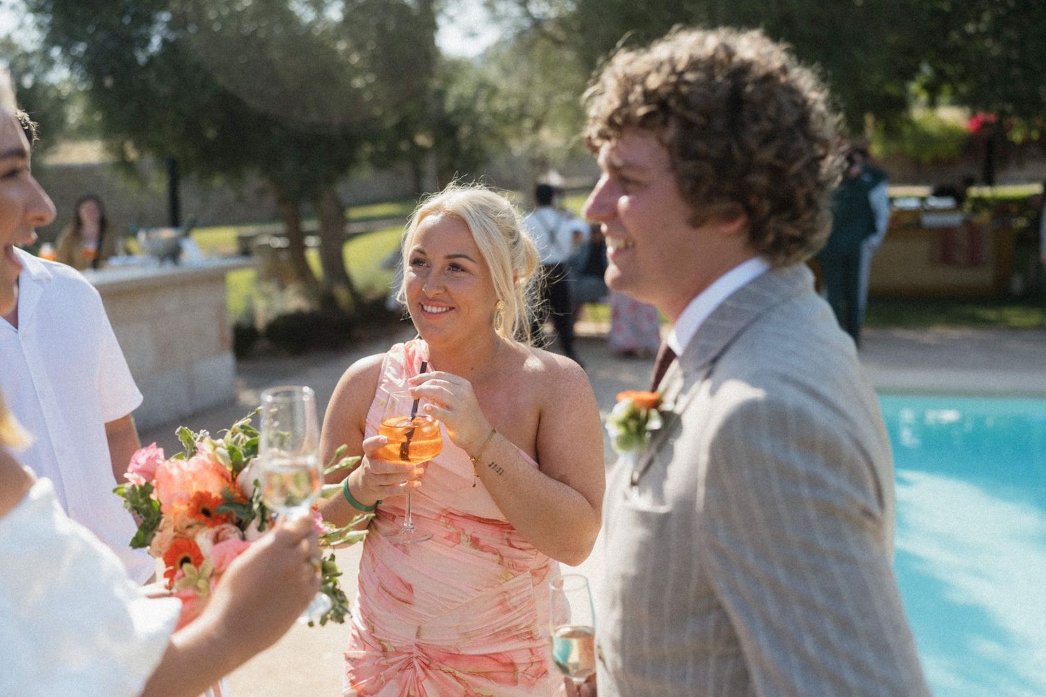 Guests in formal attire socializing outdoors near a pool at a sunny wedding reception.