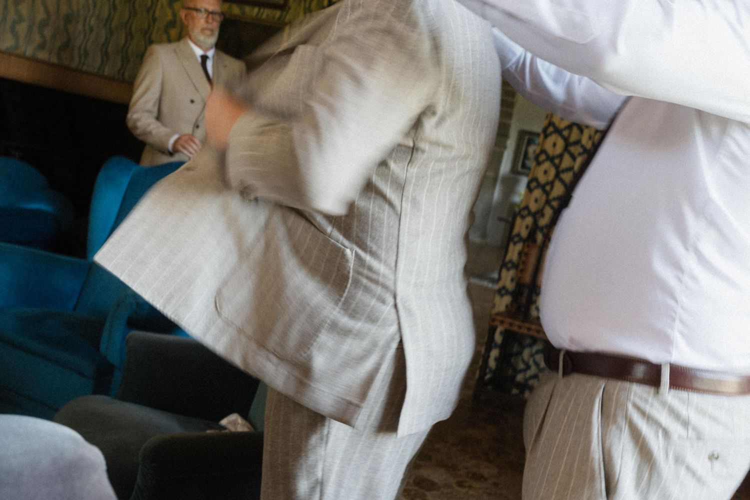 Two men in suits having a pillow fight while an older man in glasses watches disapprovingly in the background.