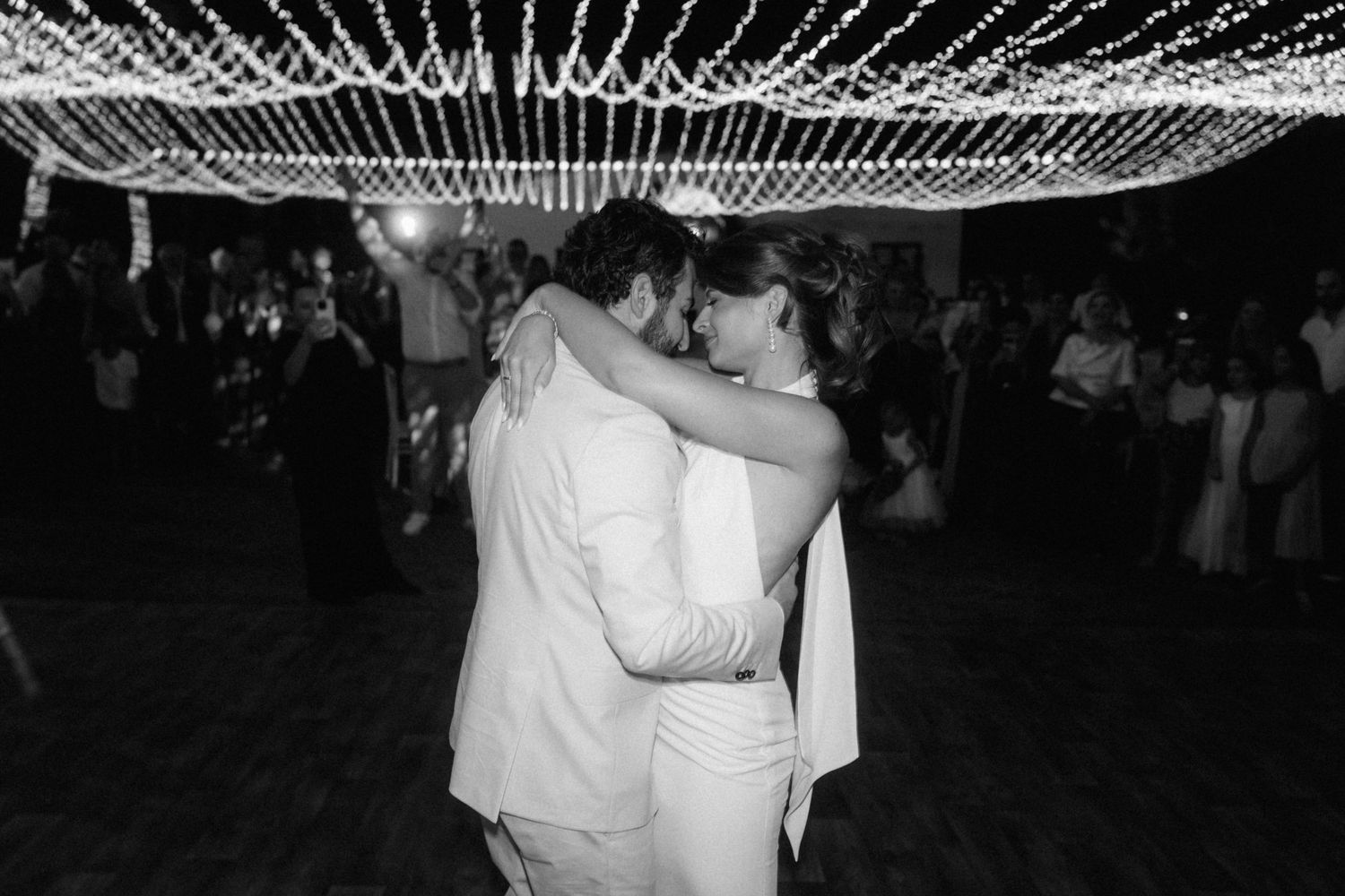 Black and white photo of bride and groom sharing first dance under twinkling string lights at wedding reception.