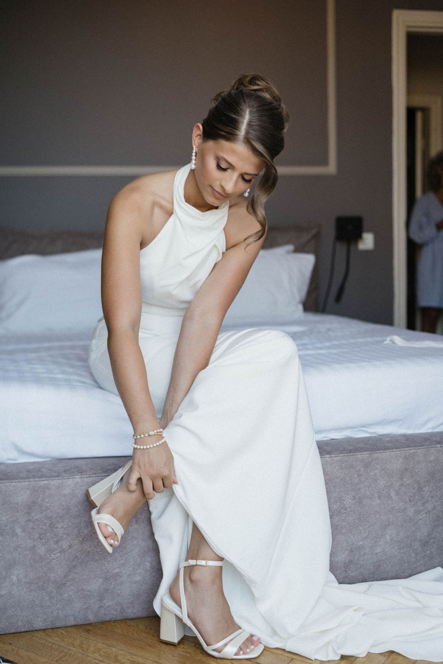 Bride in white halter wedding dress putting on strappy heels while sitting on bed before ceremony.