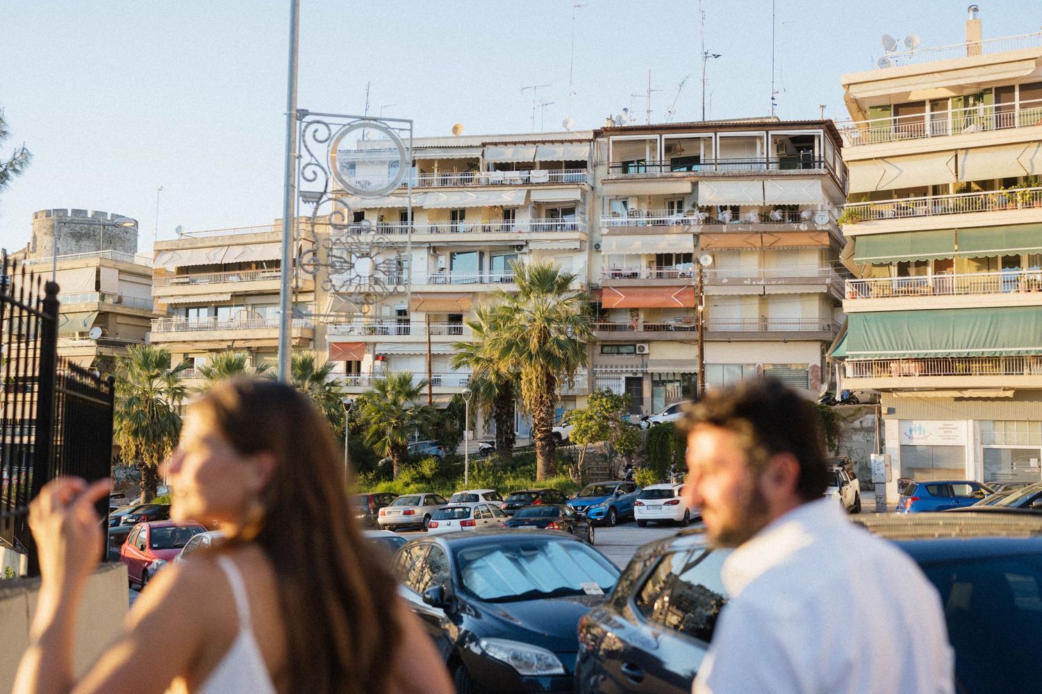 Couple in foreground with Mediterranean apartment buildings, palm trees, and parked cars in sunny urban setting.