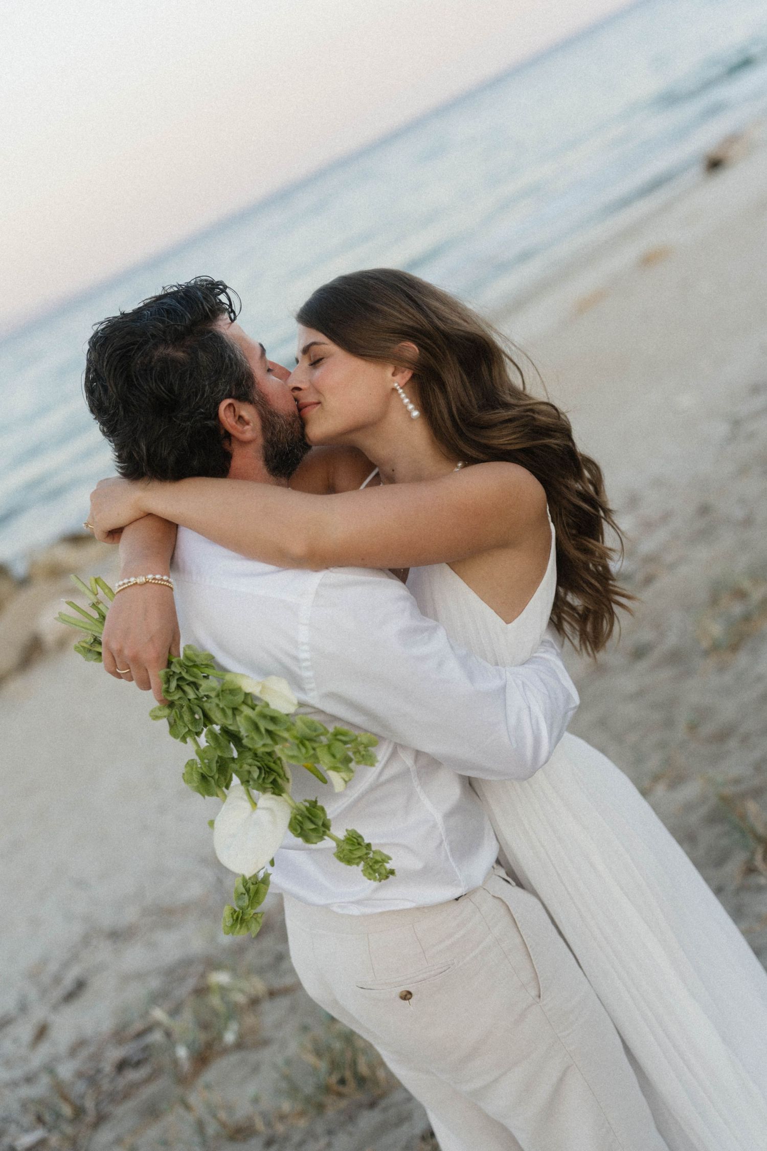 Groom lifting bride in white dress as they kiss on rocky beach with ocean in background at sunset.