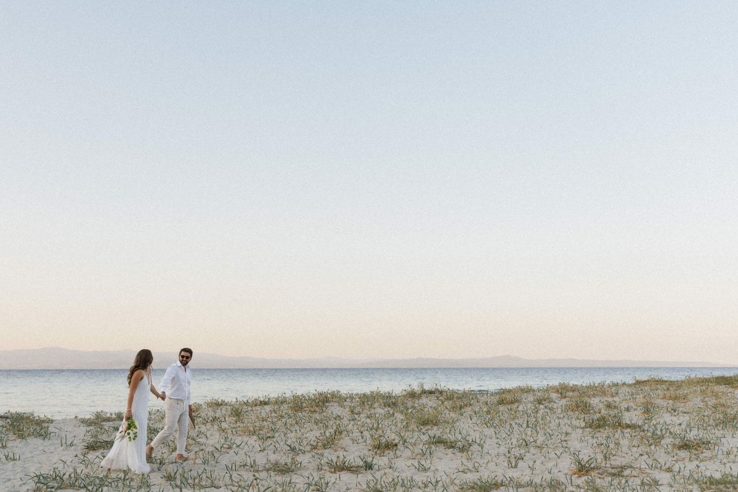 Couple in white attire holds hands on a serene beach at dusk with calm ocean and pastel sky backdrop.