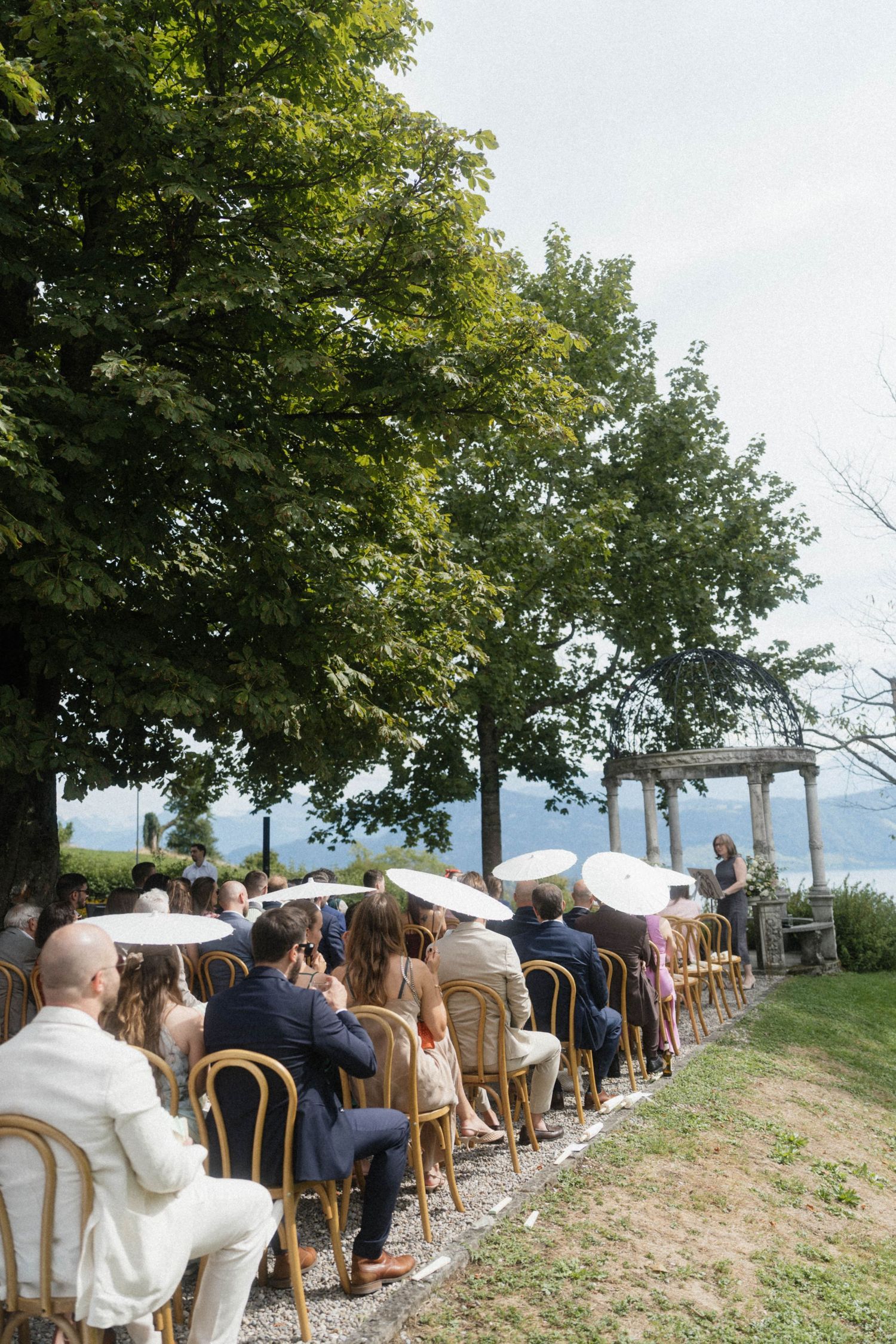 Wedding guests seated outdoors under trees with lake views during ceremony.