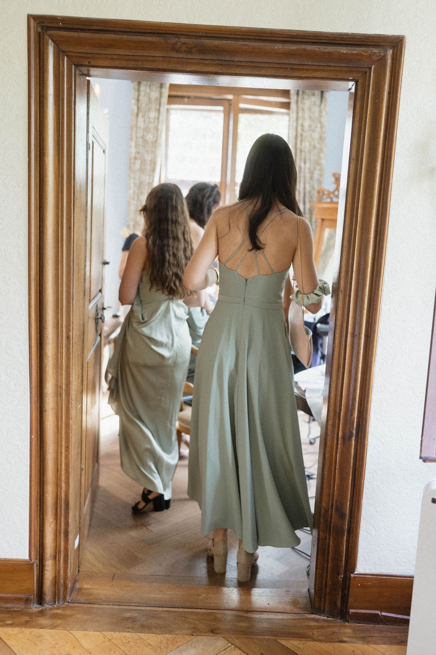 Two bridesmaids in sage green dresses walk through an elegant wooden doorway during wedding preparations.