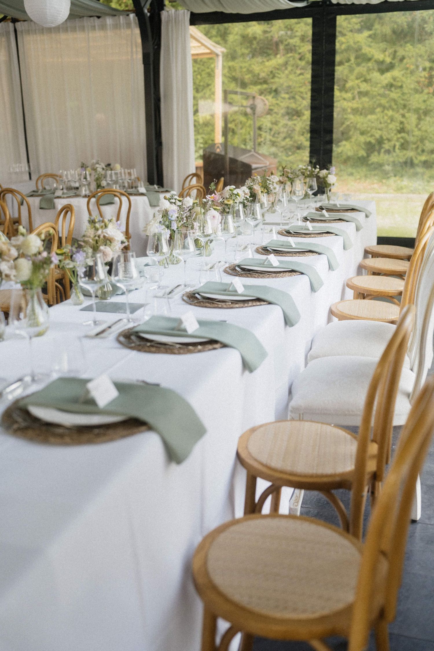 Elegantly set wedding reception table with sage green napkins, wicker chargers, and floral centerpieces under a tent.