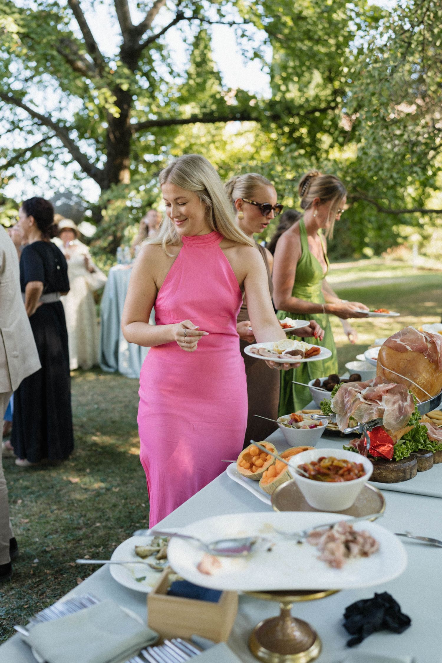 Woman in pink dress serving herself at an outdoor garden party buffet table with guests in background.