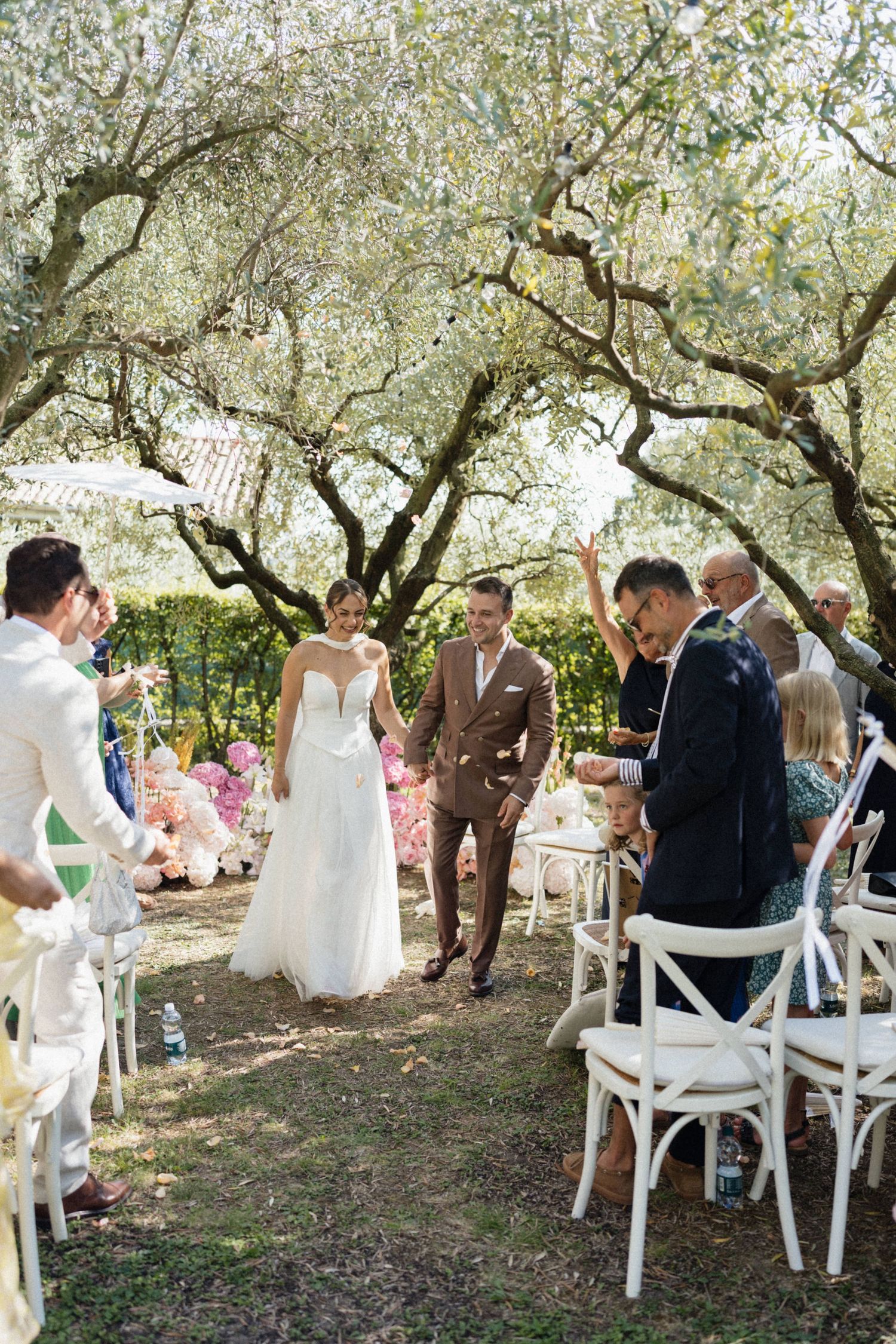 Bride and groom walk down the aisle at an outdoor garden wedding ceremony under olive trees.
