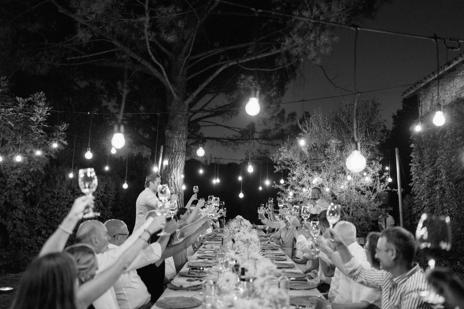 Guests raise glasses in a toast at an outdoor wedding reception under warm string lights at night.