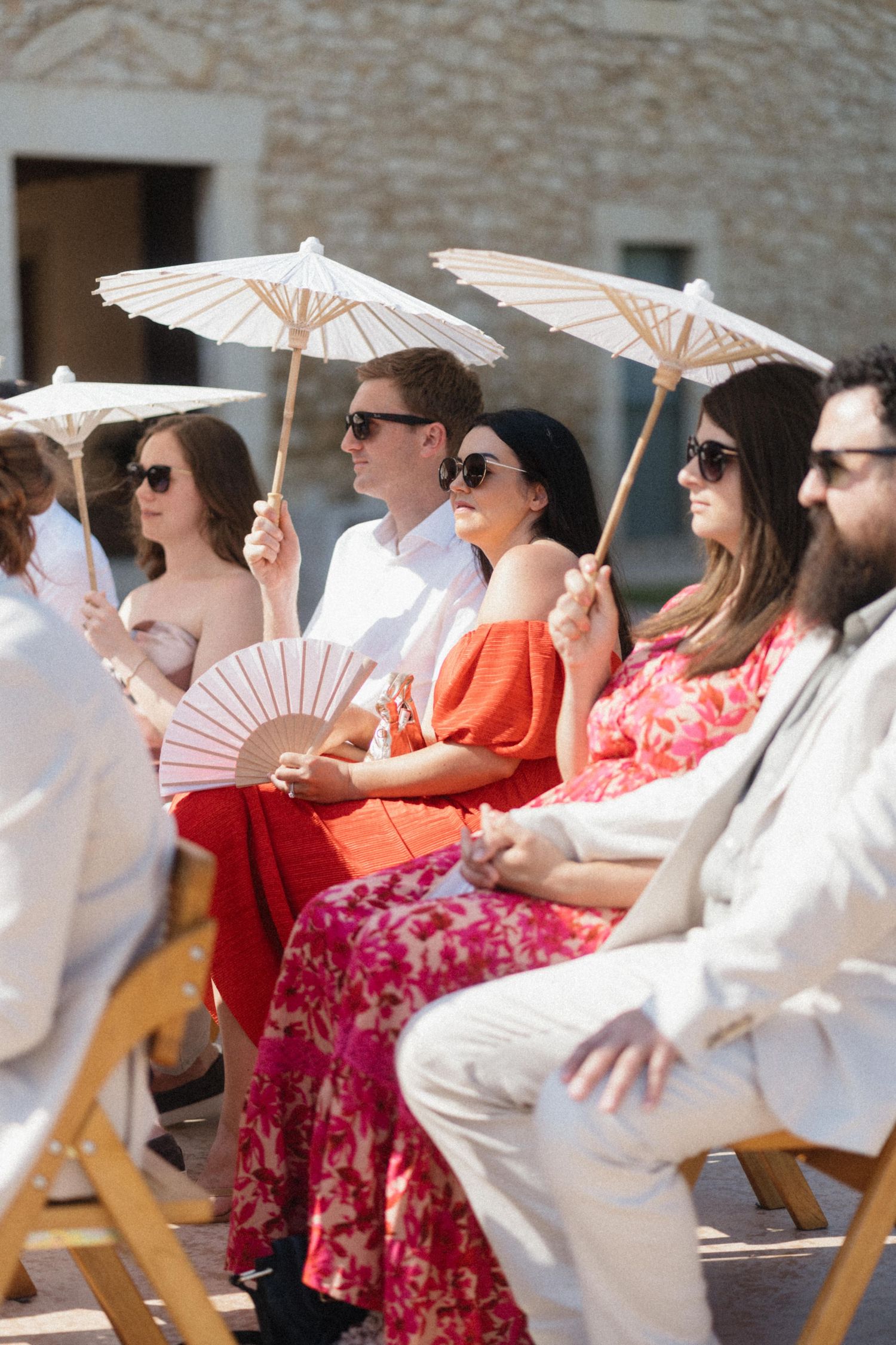 Wedding guests in colorful dresses hold white parasols and fans while seated outdoors at a sunny ceremony.