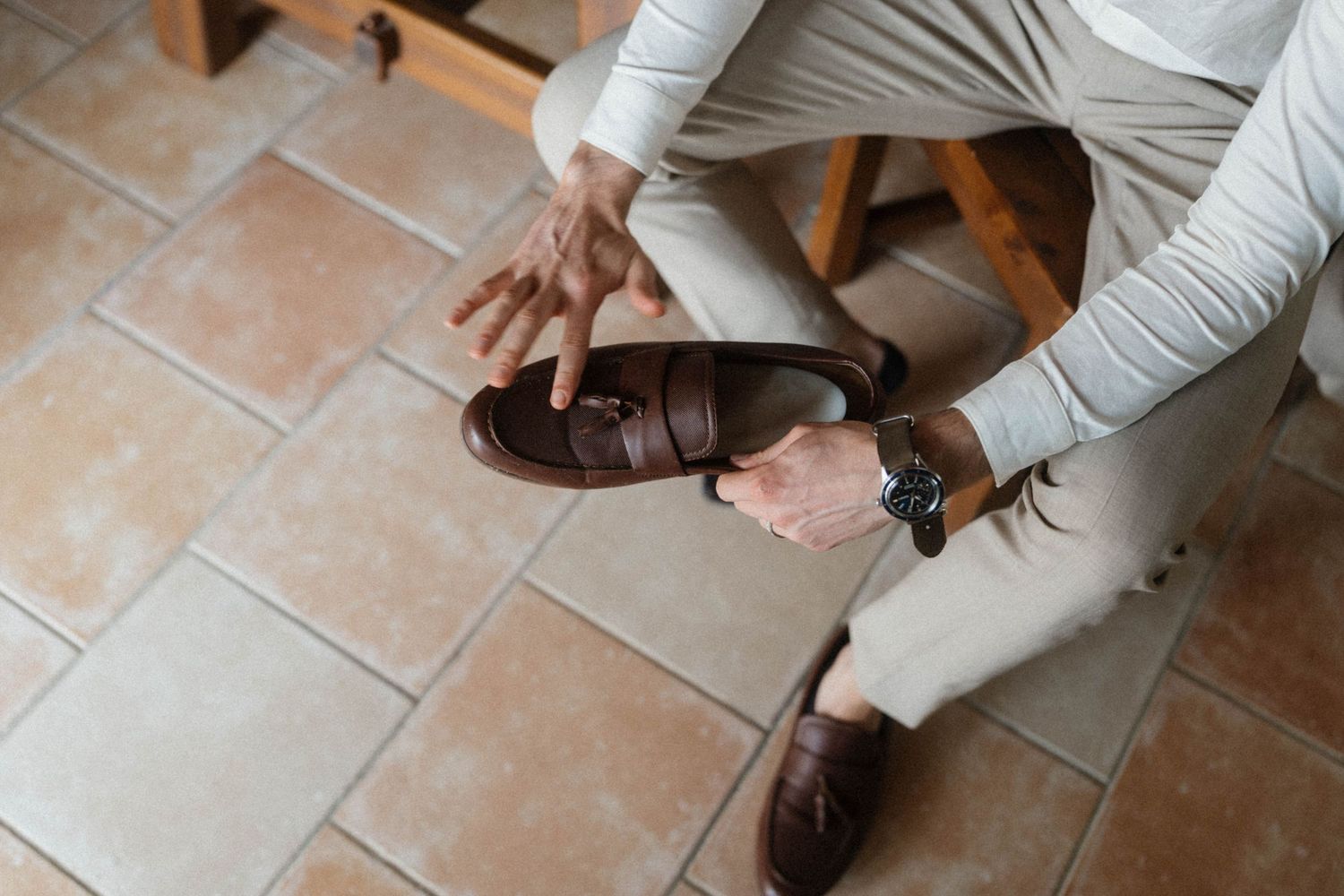 Person holding a brown leather loafer shoe while seated, wearing beige trousers and a watch on tiled floor.
